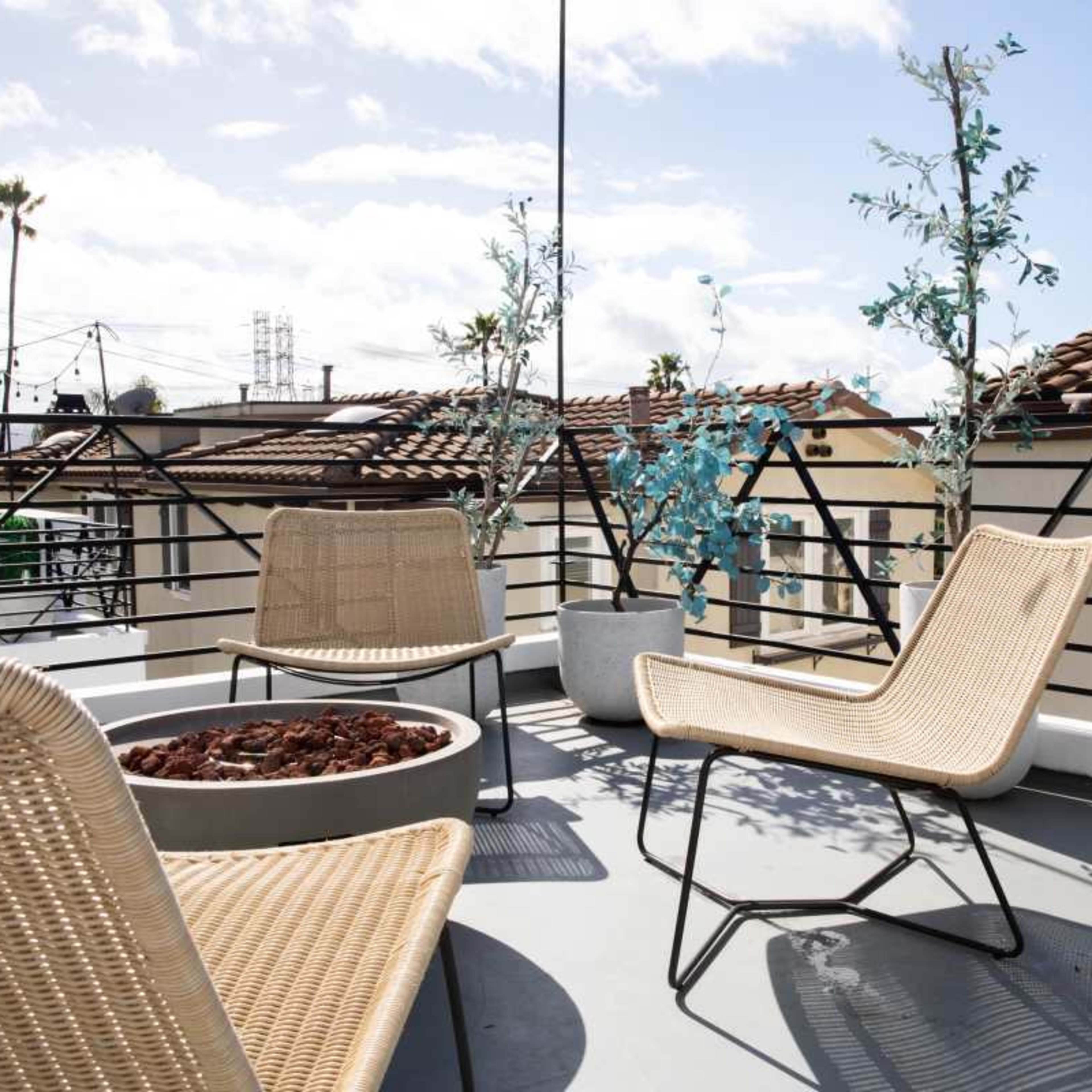 A rooftop terrace with three woven chairs, a round fire pit surrounded by small rocks, and two potted trees against a backdrop of a blue sky.