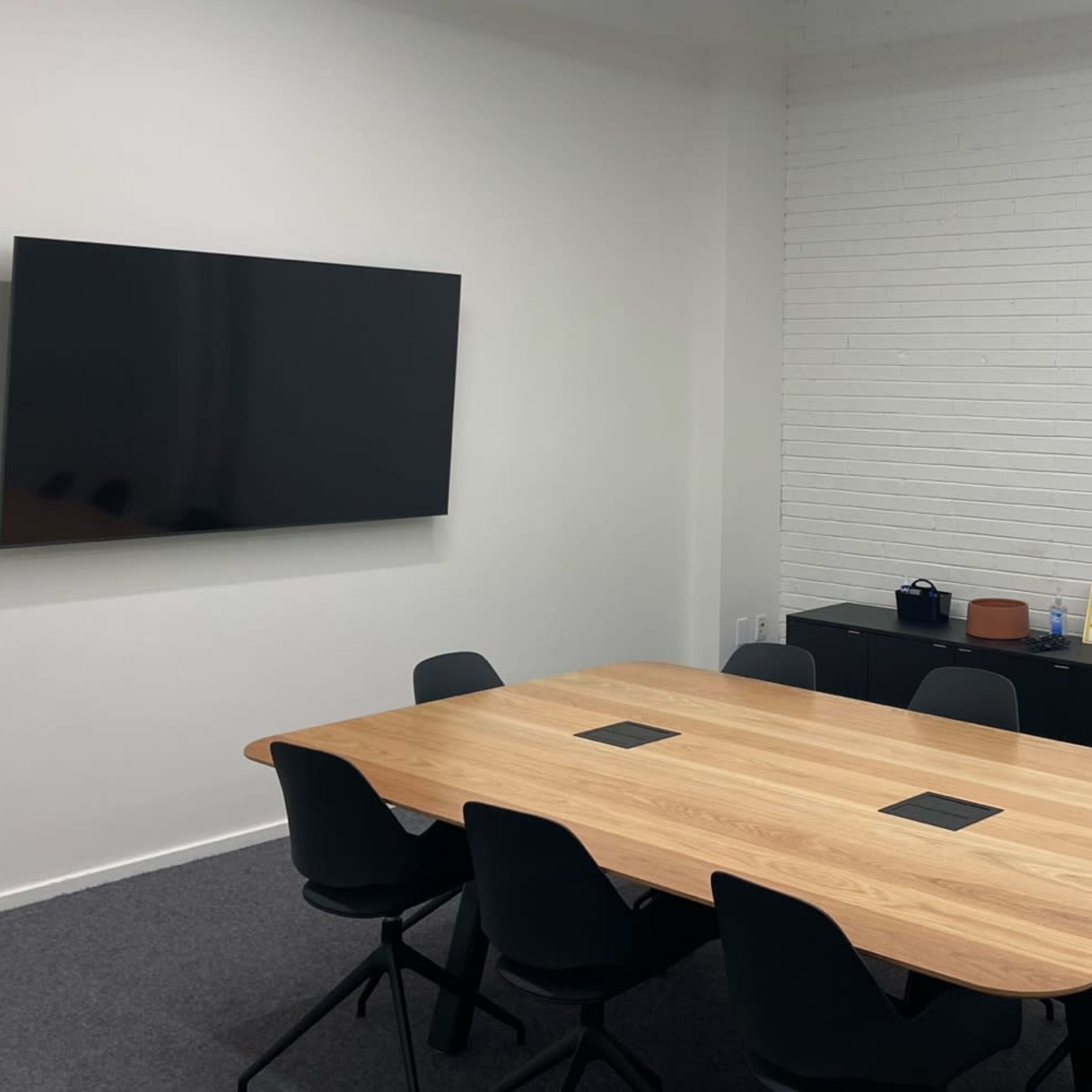 A meeting room features a long wooden table surrounded by black chairs, with a wall-mounted television and a low black cabinet.