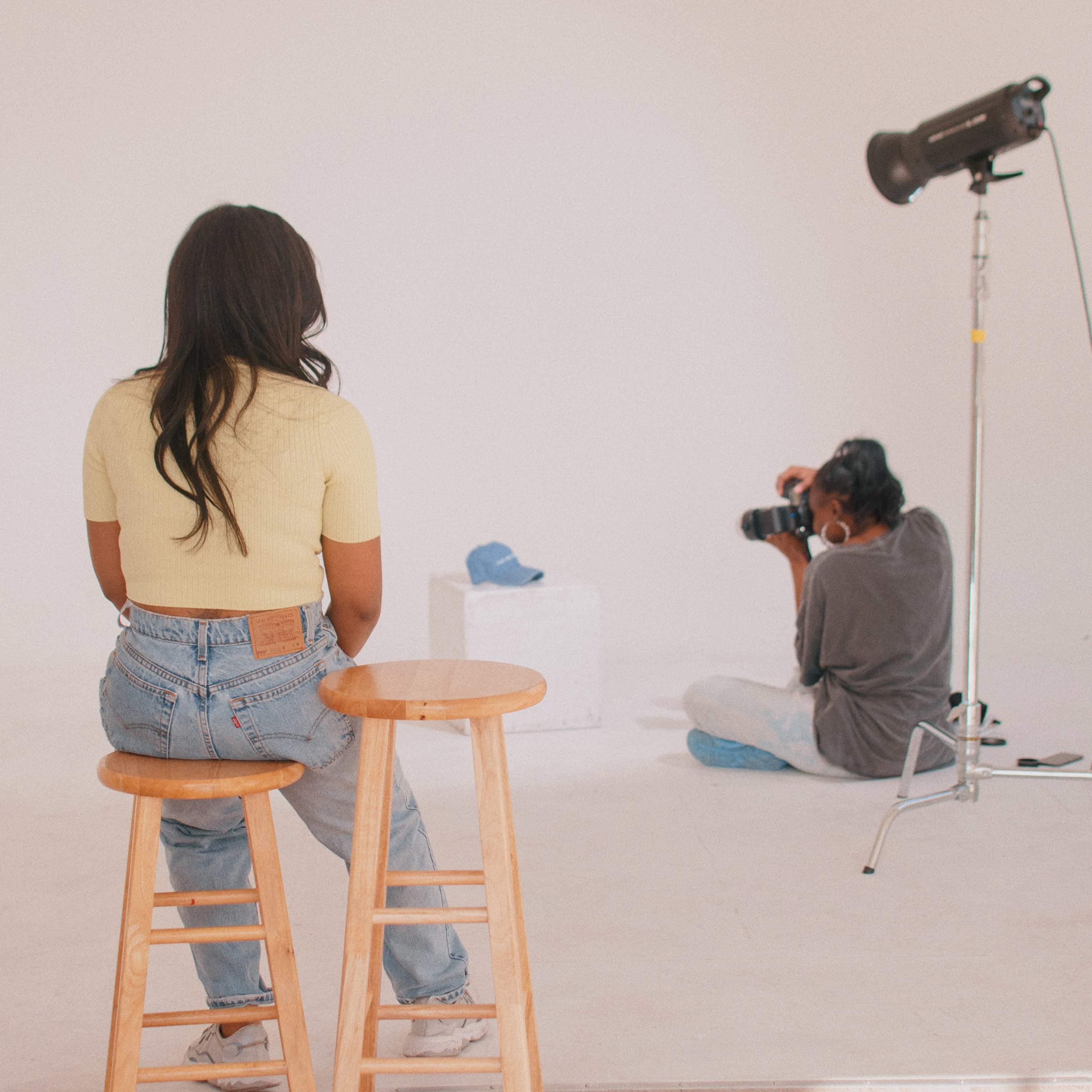A woman sits on a stool in a studio while a photographer captures an image of a blue shoe on a pedestal.