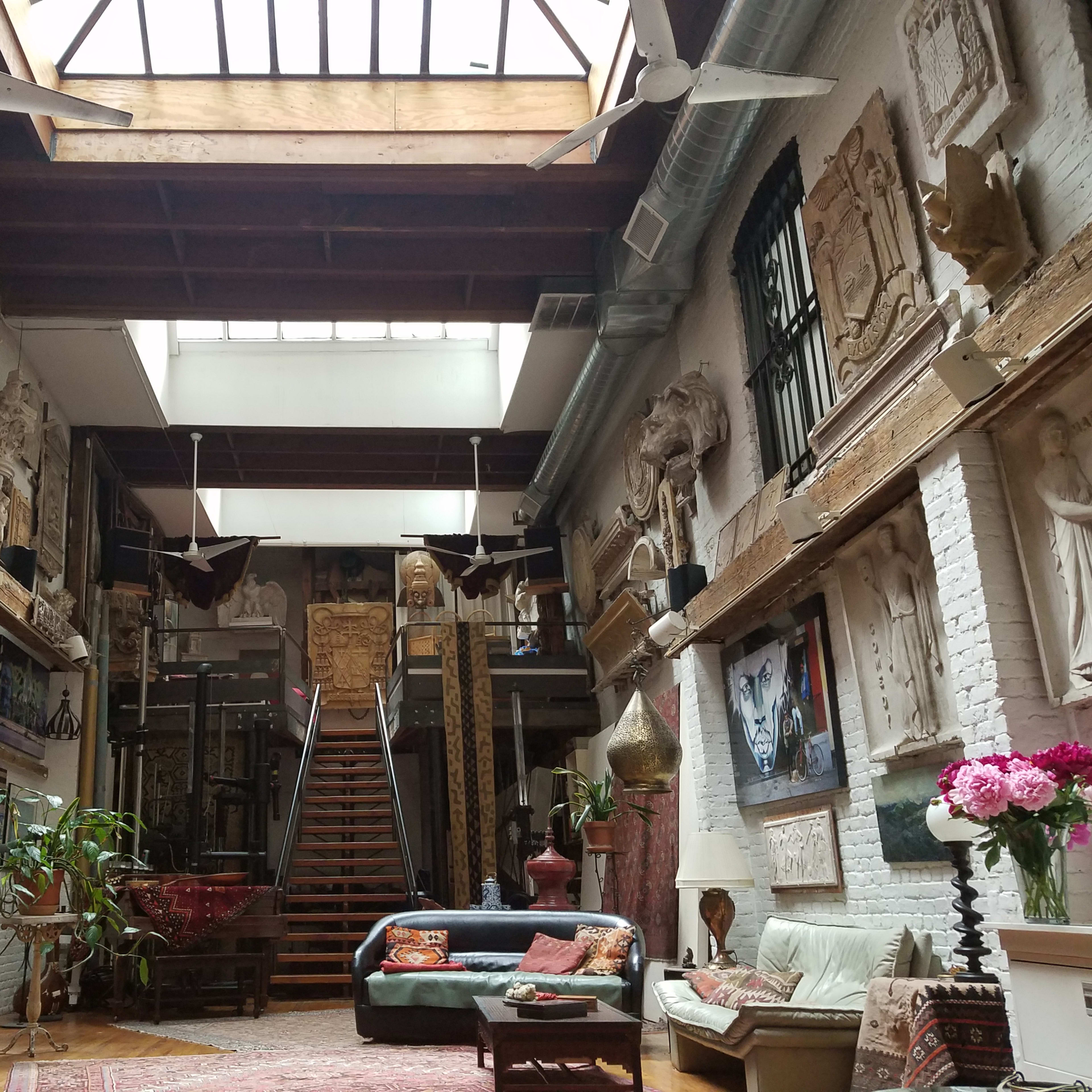 A spacious, eclectic living area featuring high ceilings, numerous sculptures and wall art, and a staircase leading to an upper level.
