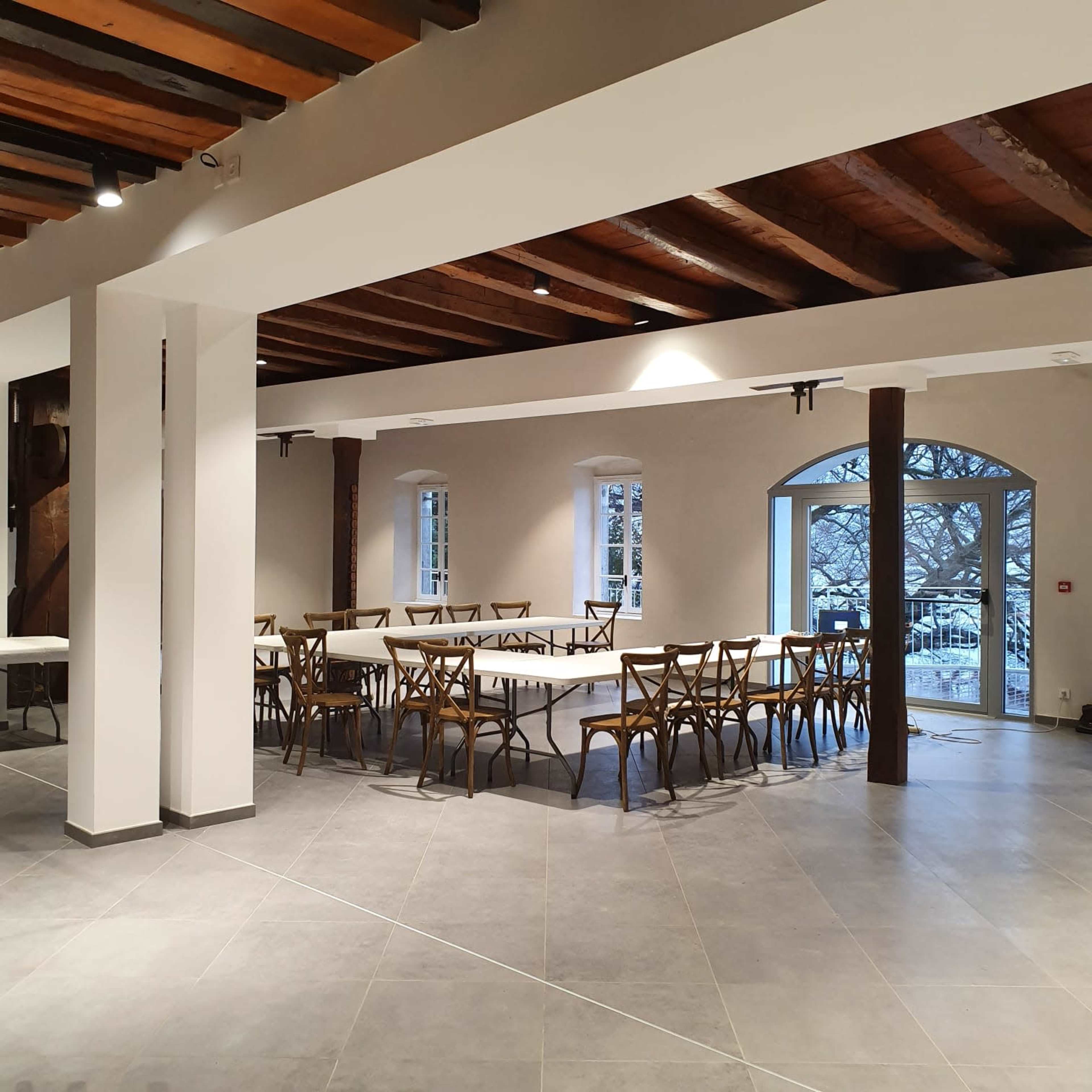 An empty indoor space with several rectangular tables arranged for seating, large windows letting in natural light, and wooden beams on the ceiling.