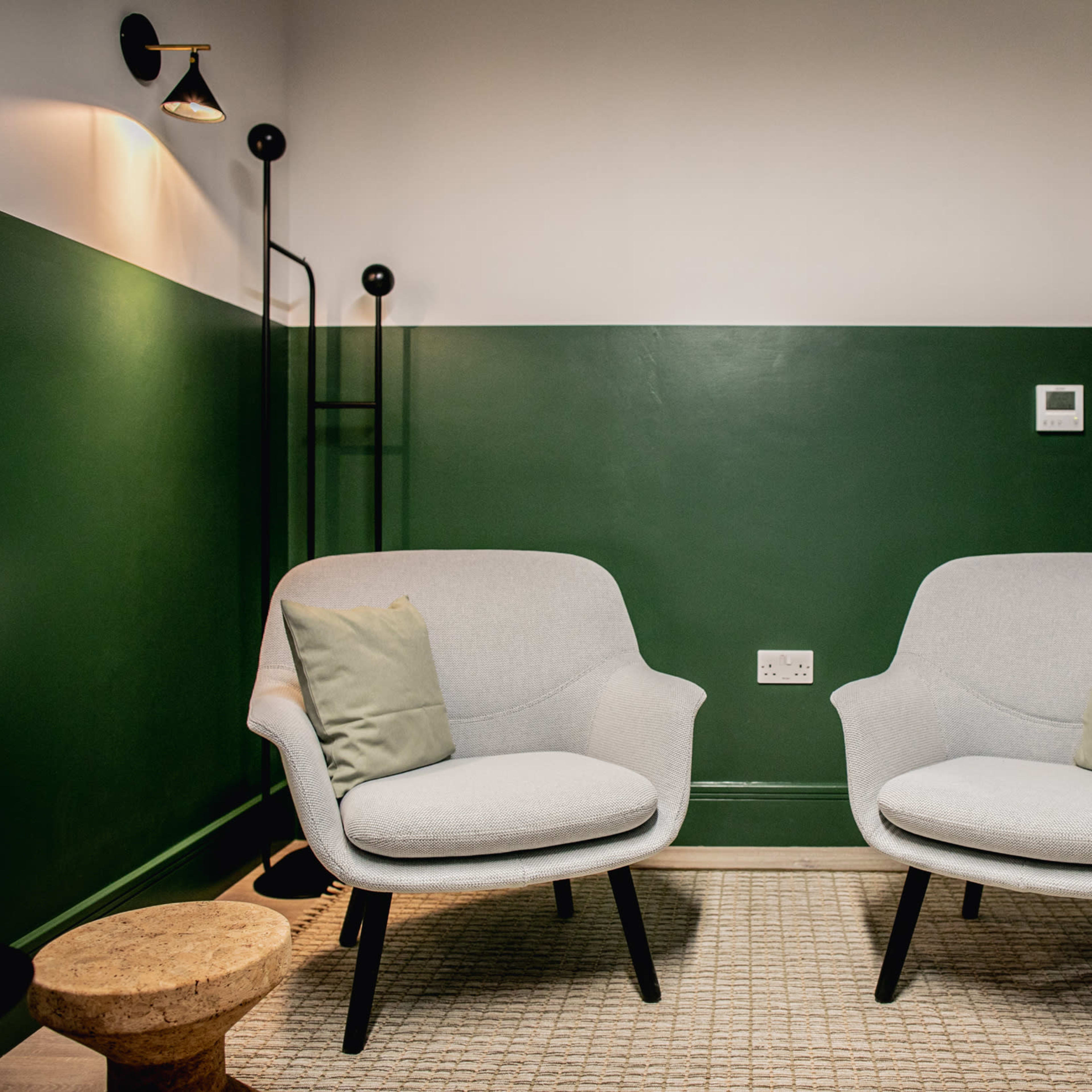 Two light gray chairs with green cushions are positioned opposite each other in a room with a dark green accent wall, beside a small wooden stool.