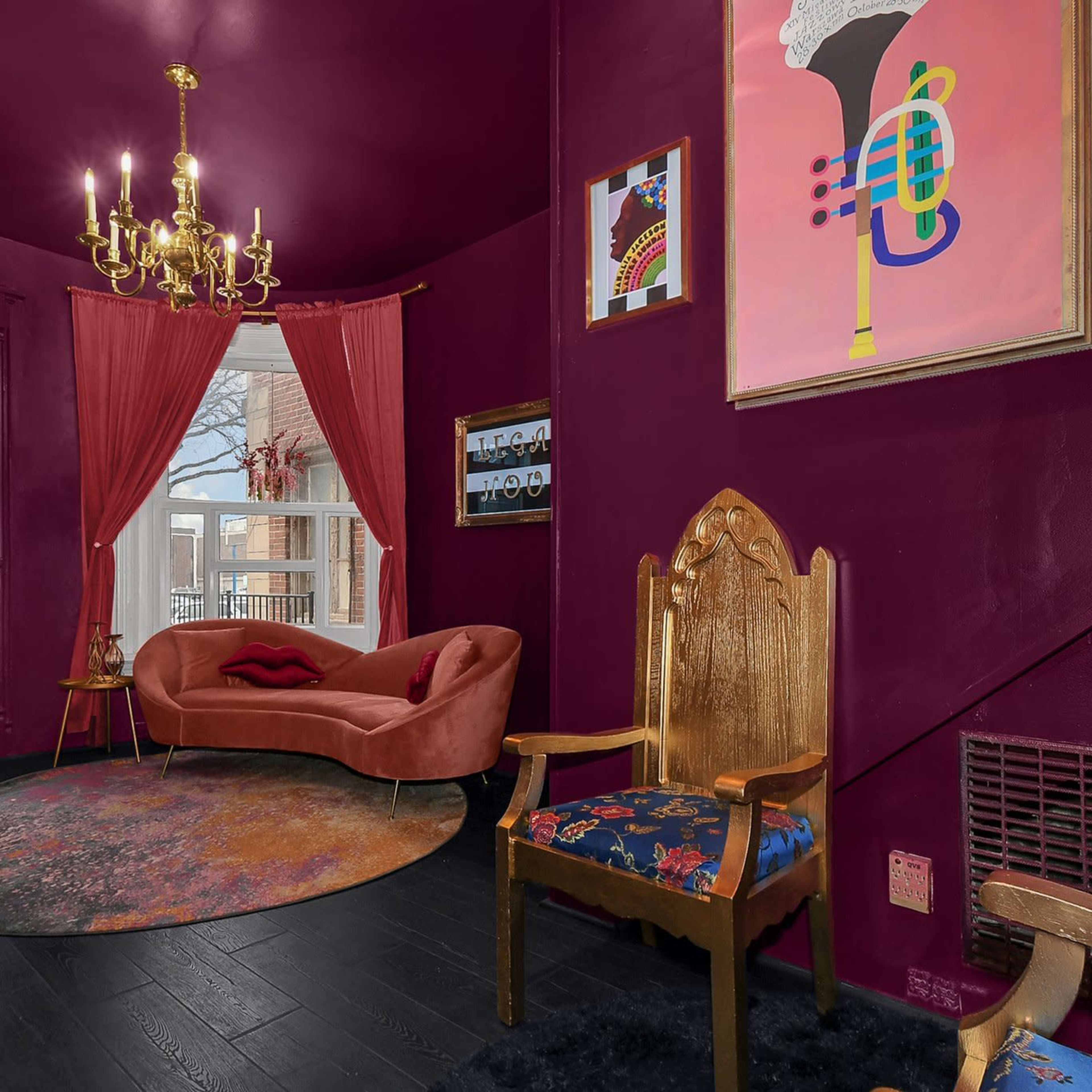 A small entryway featuring a velvet couch, a decorative gold chandelier, and a vintage armchair, all set against dark maroon walls.