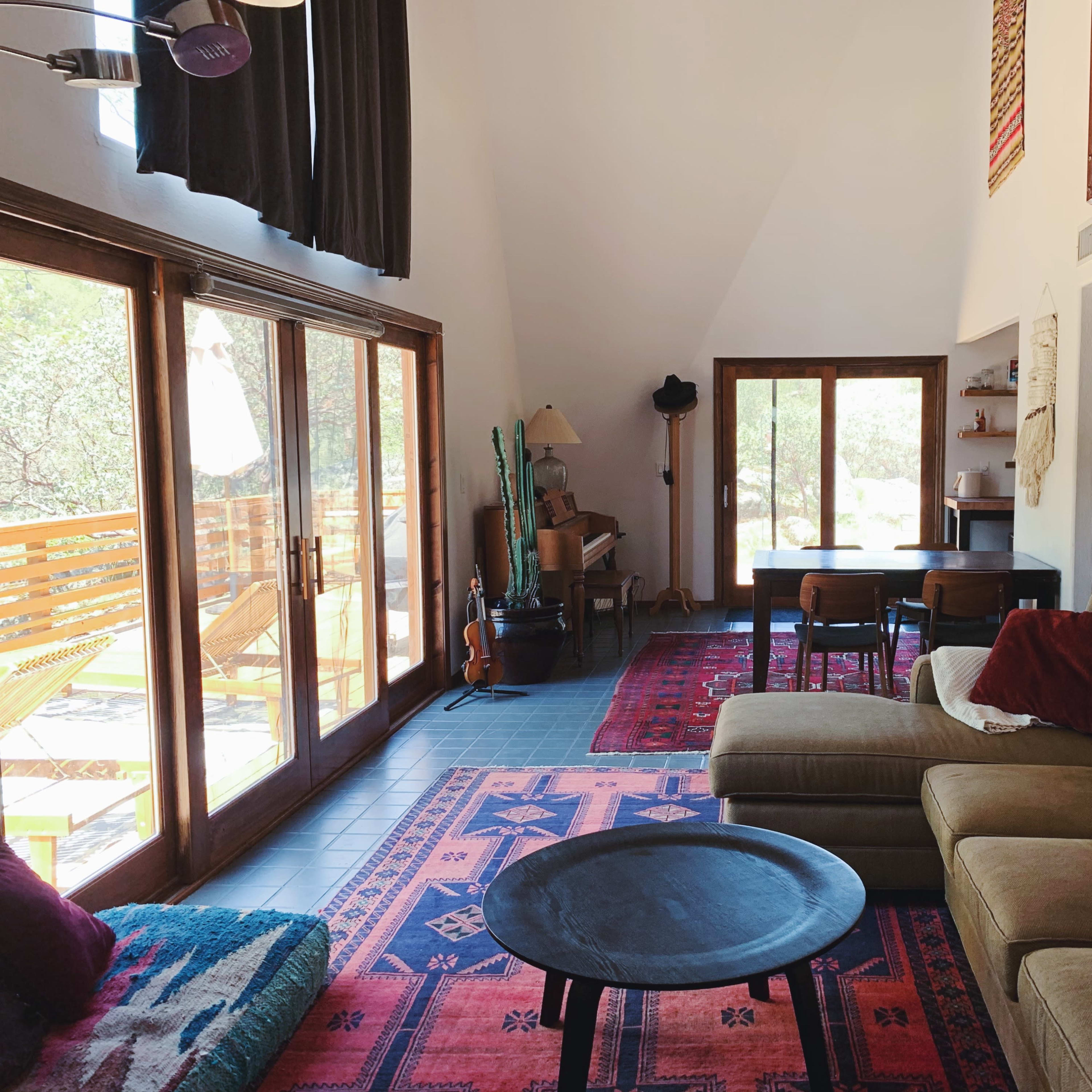 The interior of a modern living room features a large, beige sectional sofa, a round black coffee table, and large windows overlooking a patio, with wooden accents and various musical instruments displayed on the walls.