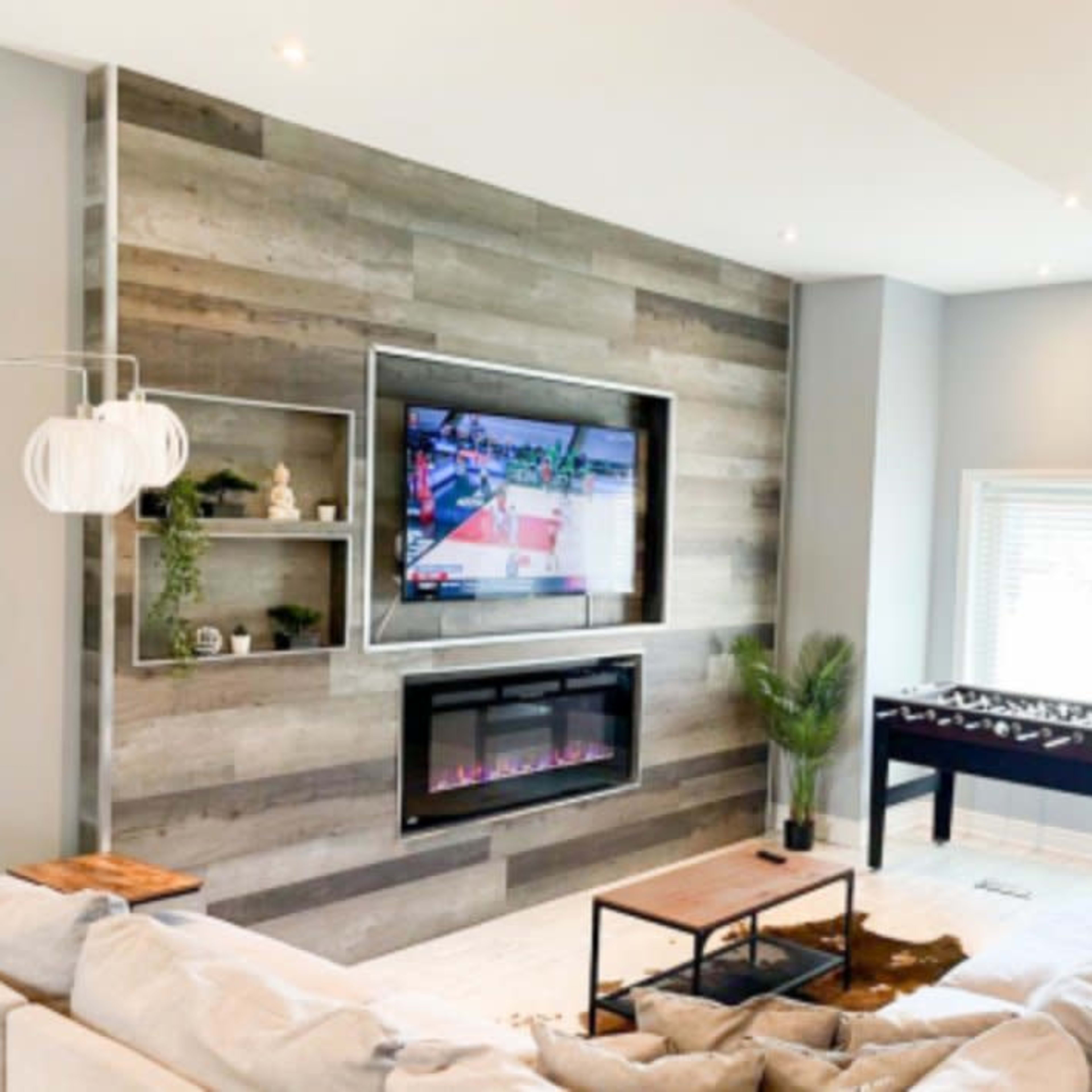 A modern living room features a wall-mounted TV above a sleek electric fireplace, flanked by decorative shelves, with a foosball table in the corner.