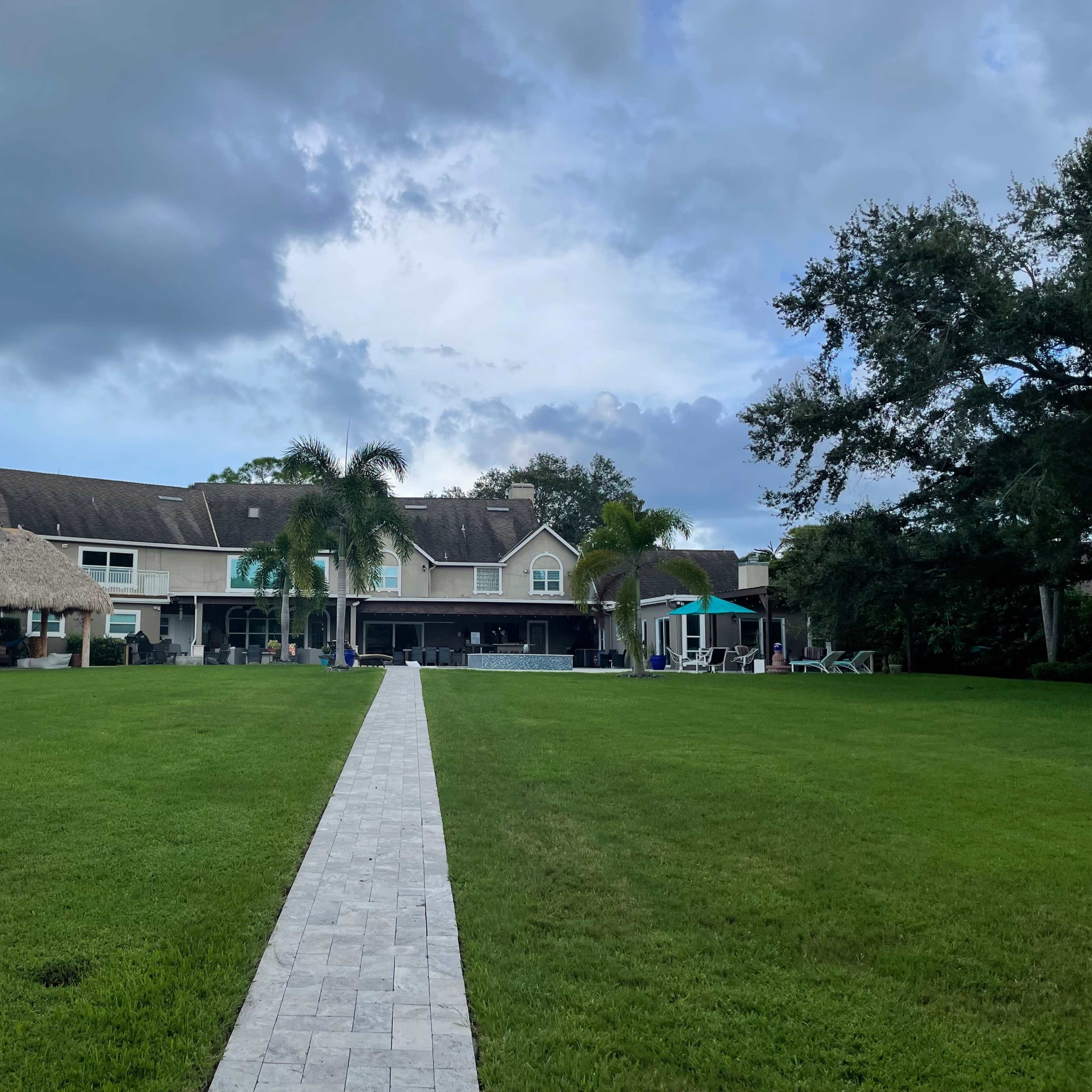 A large house with a thatched roof and a patio is set against a cloudy sky, facing a well-maintained lawn and a stone pathway.