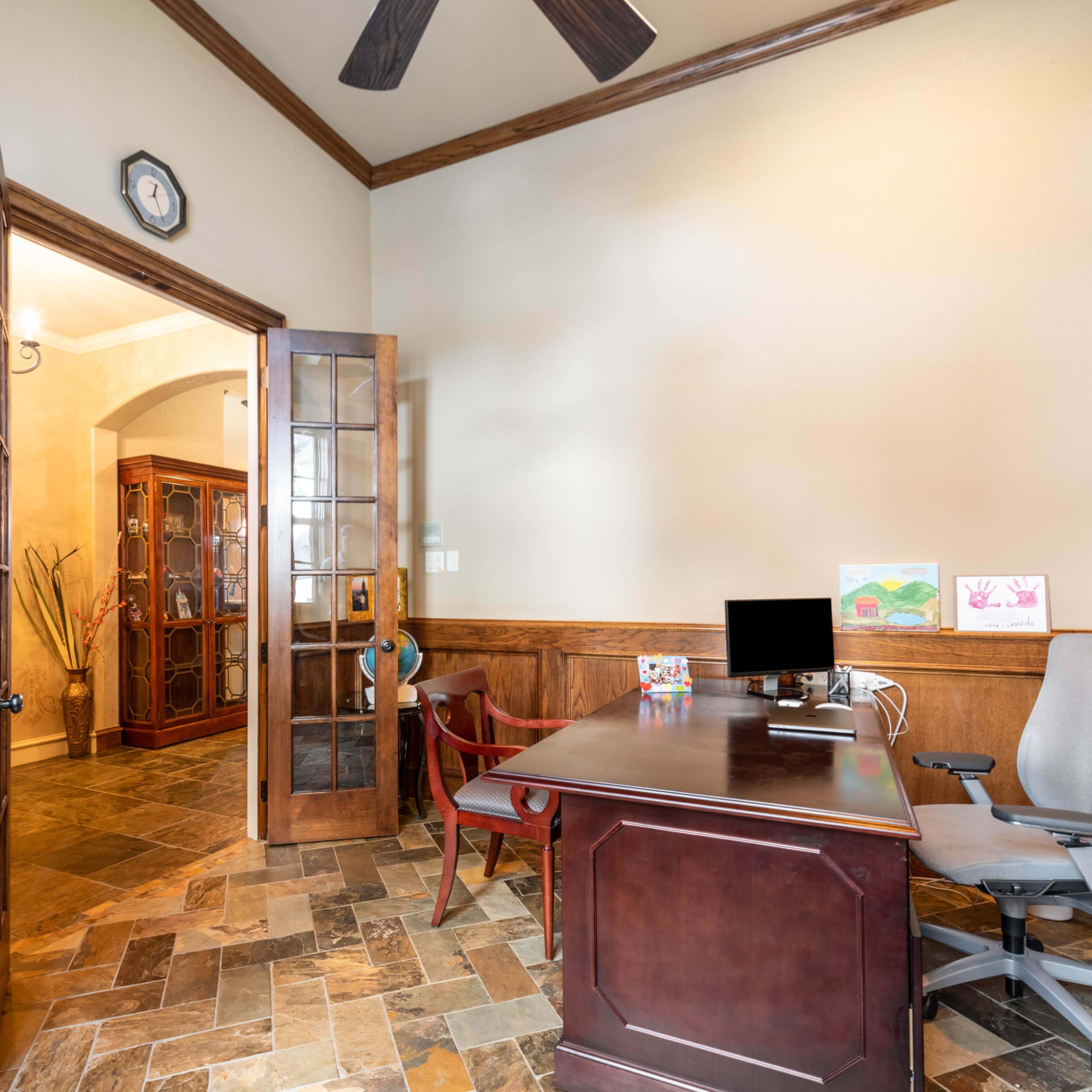 An office space with a wooden desk, office chair, and a doorway leading to a room with a glass cabinet and art displayed on a wall.