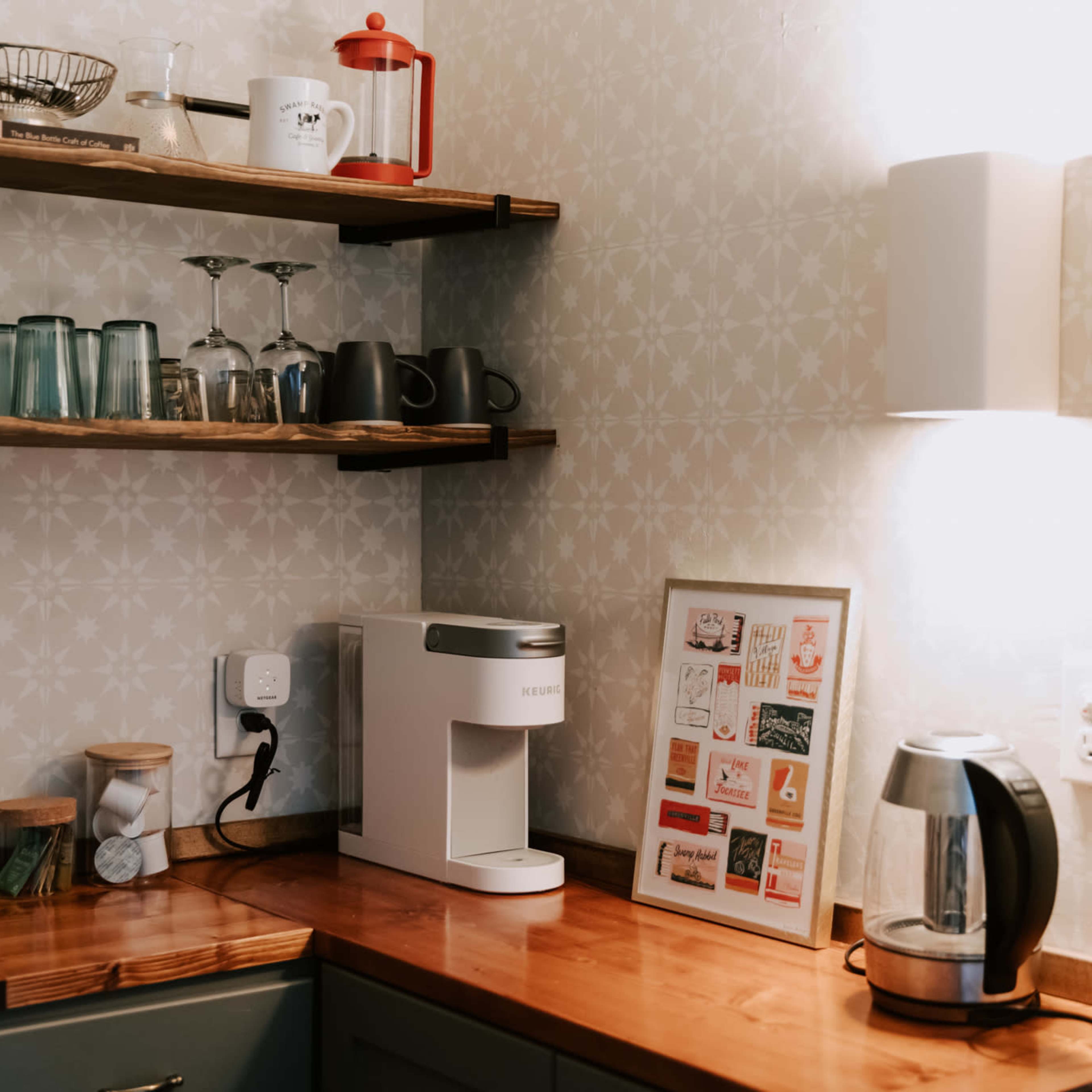 A kitchen corner features wooden shelves holding glassware and a coffee press above a countertop with a coffee machine and electric kettle.