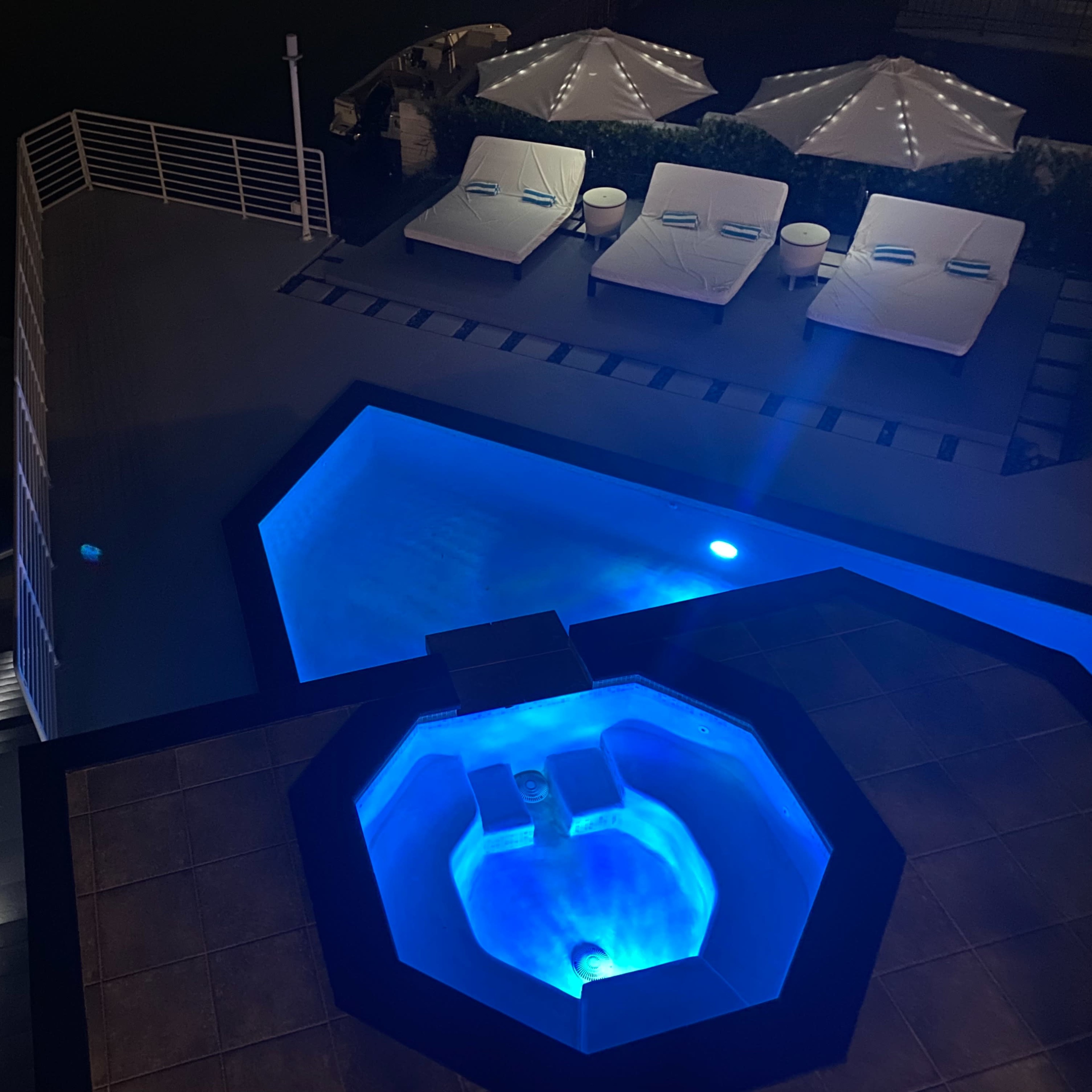 A rooftop area featuring an illuminated swimming pool, a hot tub, and lounge chairs under canopies.