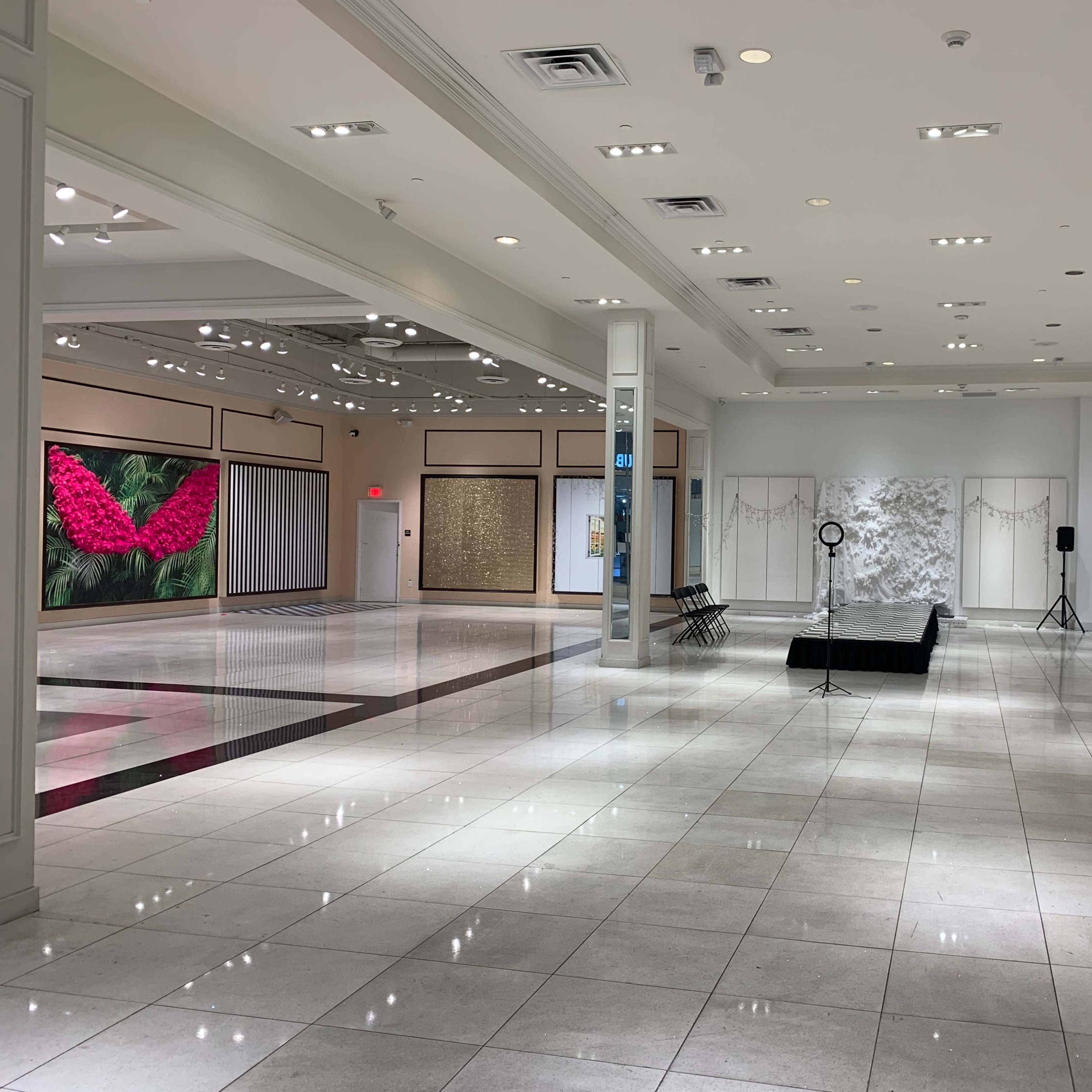 A spacious, well-lit exhibition hall featuring several empty chairs and art displays, with a large floral artwork on one wall.