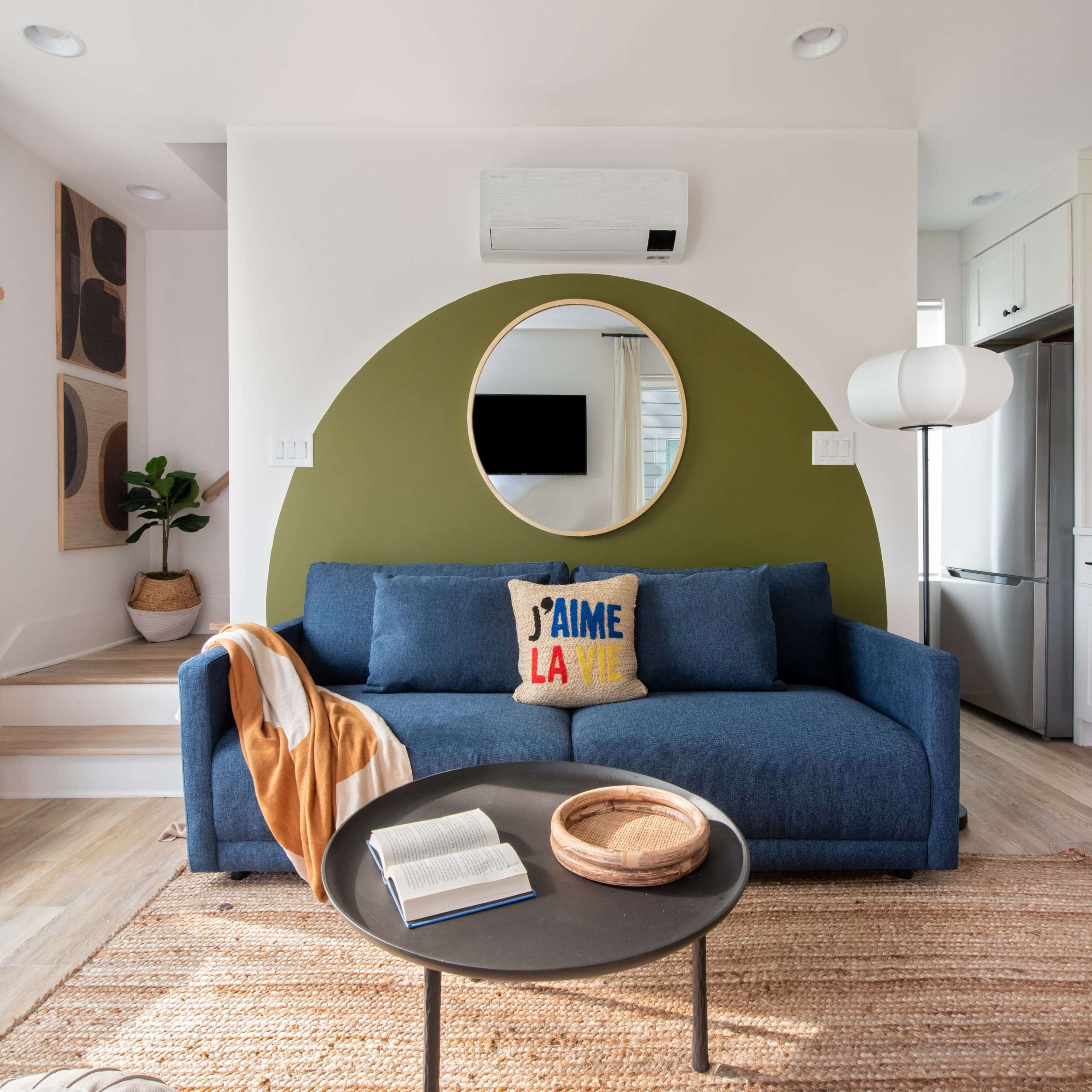 A cozy living room features a blue couch with a decorative pillow, a round mirror on a green accent wall, and a small table with a book and decorative bowl.