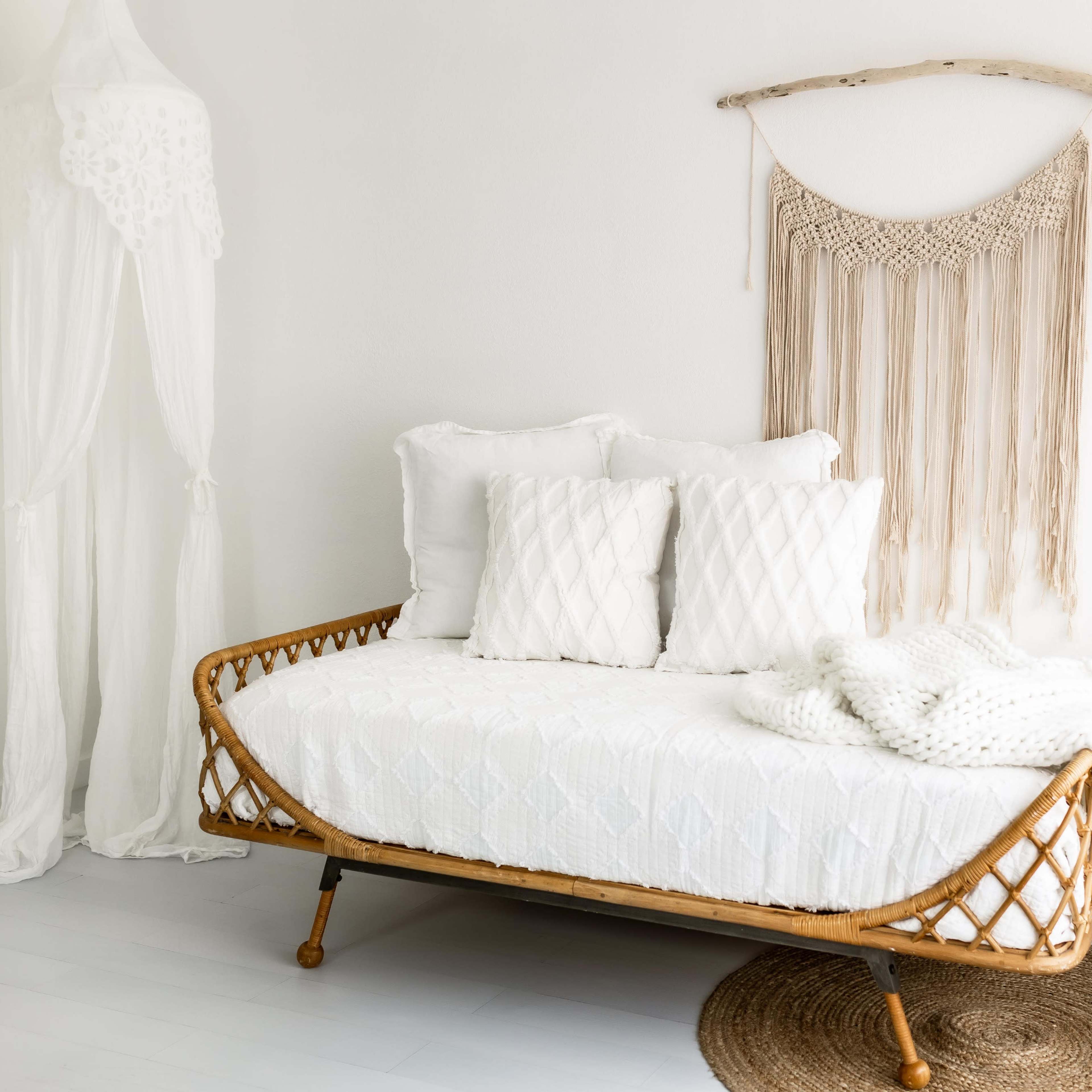 A woven daybed with white cushions is positioned against a light wall, accompanied by a macramé wall hanging, a plant in a pot, and a decorative rug.