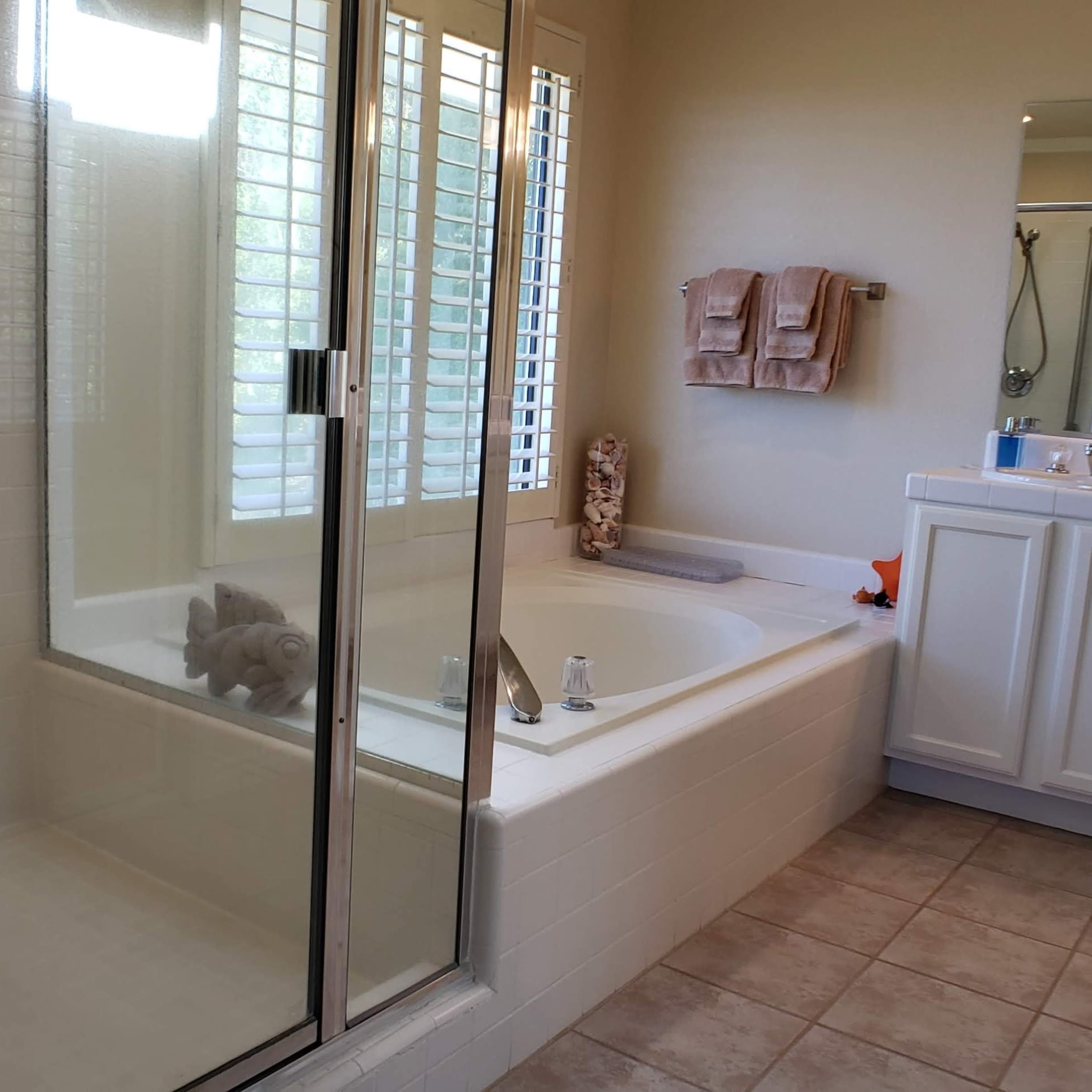 A bathroom featuring a glass-enclosed shower, a large bathtub, a double sink vanity, and beige tiled flooring.