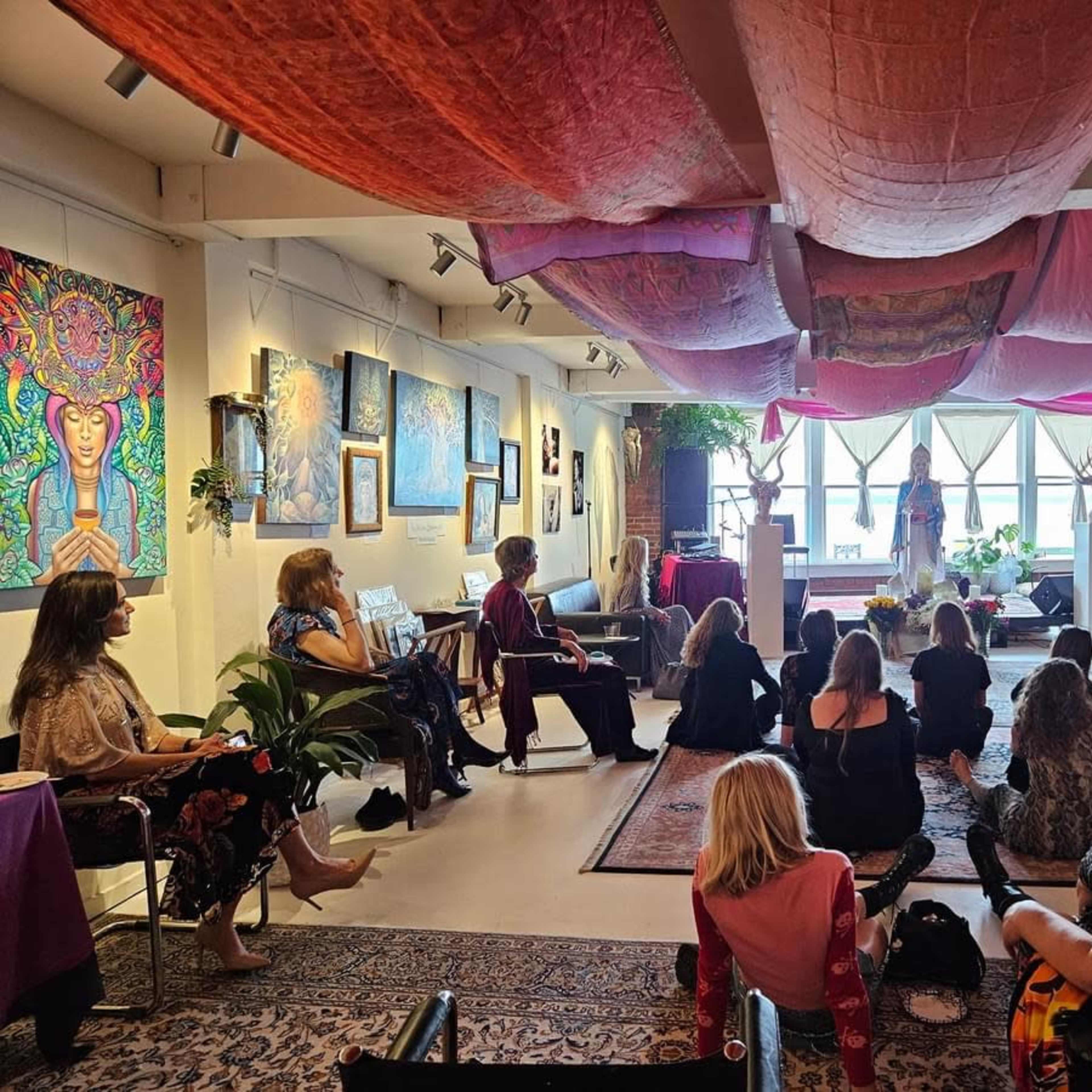 A diverse group of people sit on the floor and in chairs, listening to a speaker in a colorful, art-filled room with fabric draped from the ceiling.