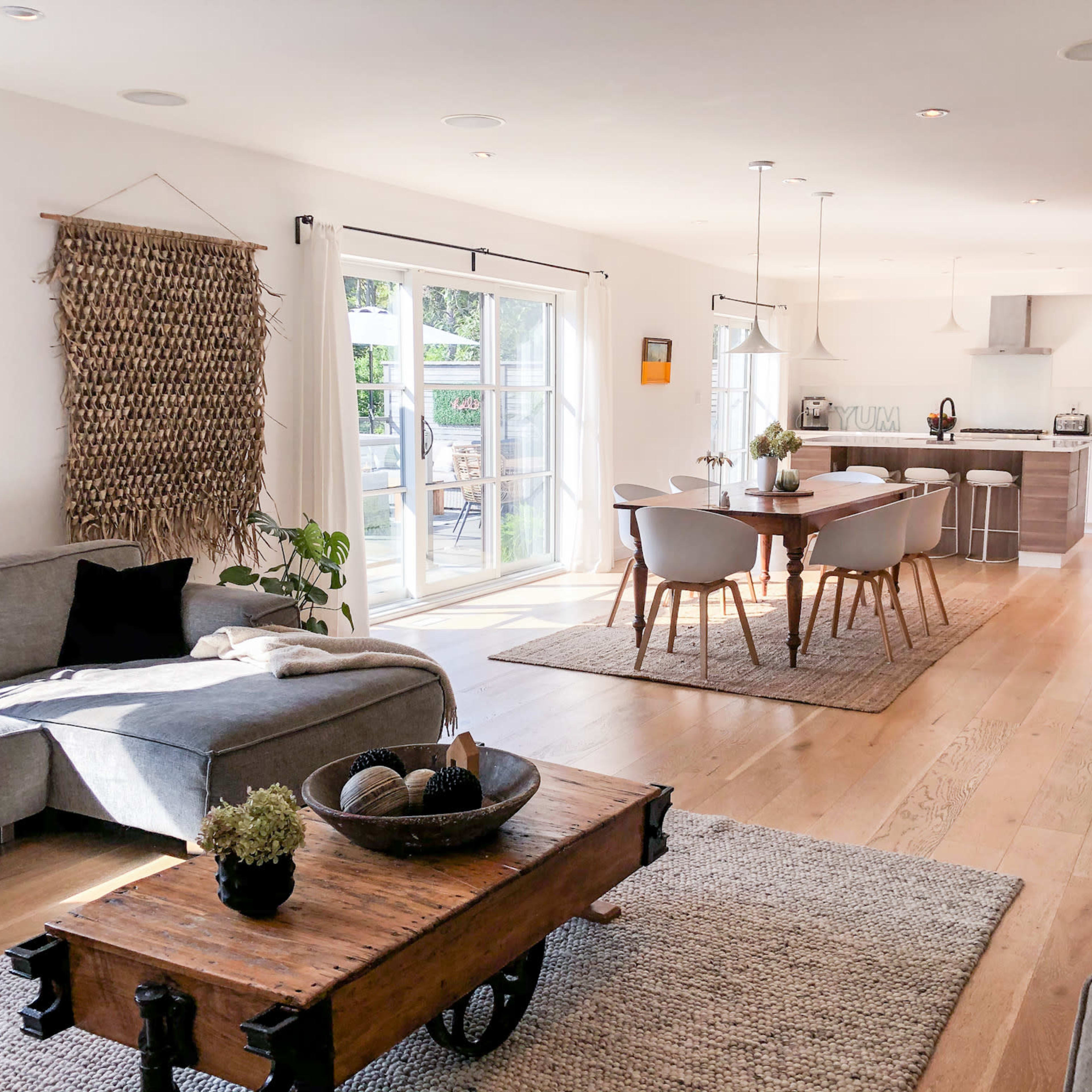 A bright, open living space featuring a gray sofa, a wooden coffee table on a textured rug, and a dining area with light wood chairs and a modern kitchen in the background.