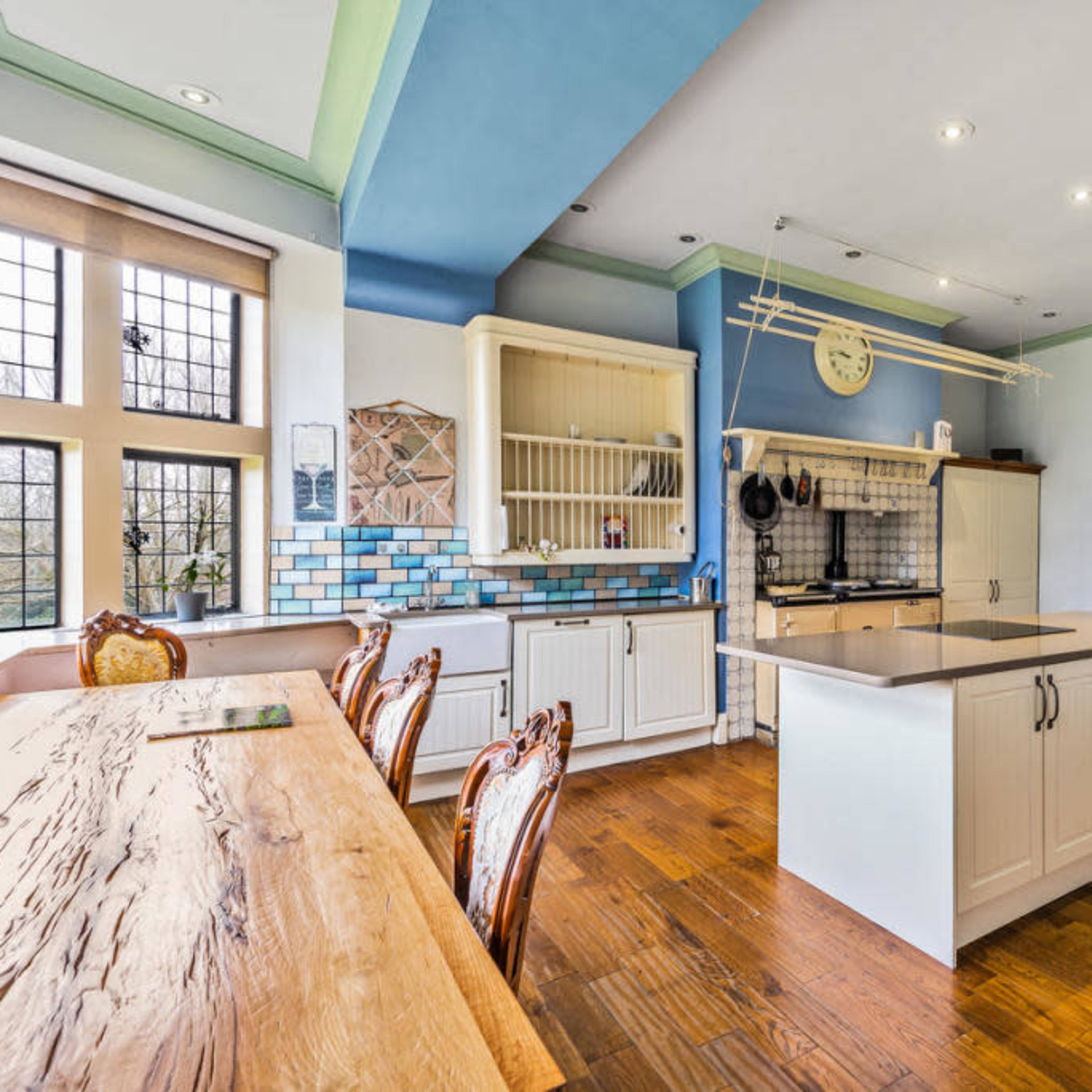 The kitchen features a large central island, wooden flooring, and blue tile accents on the walls, with a dining table and ornate chairs nearby.