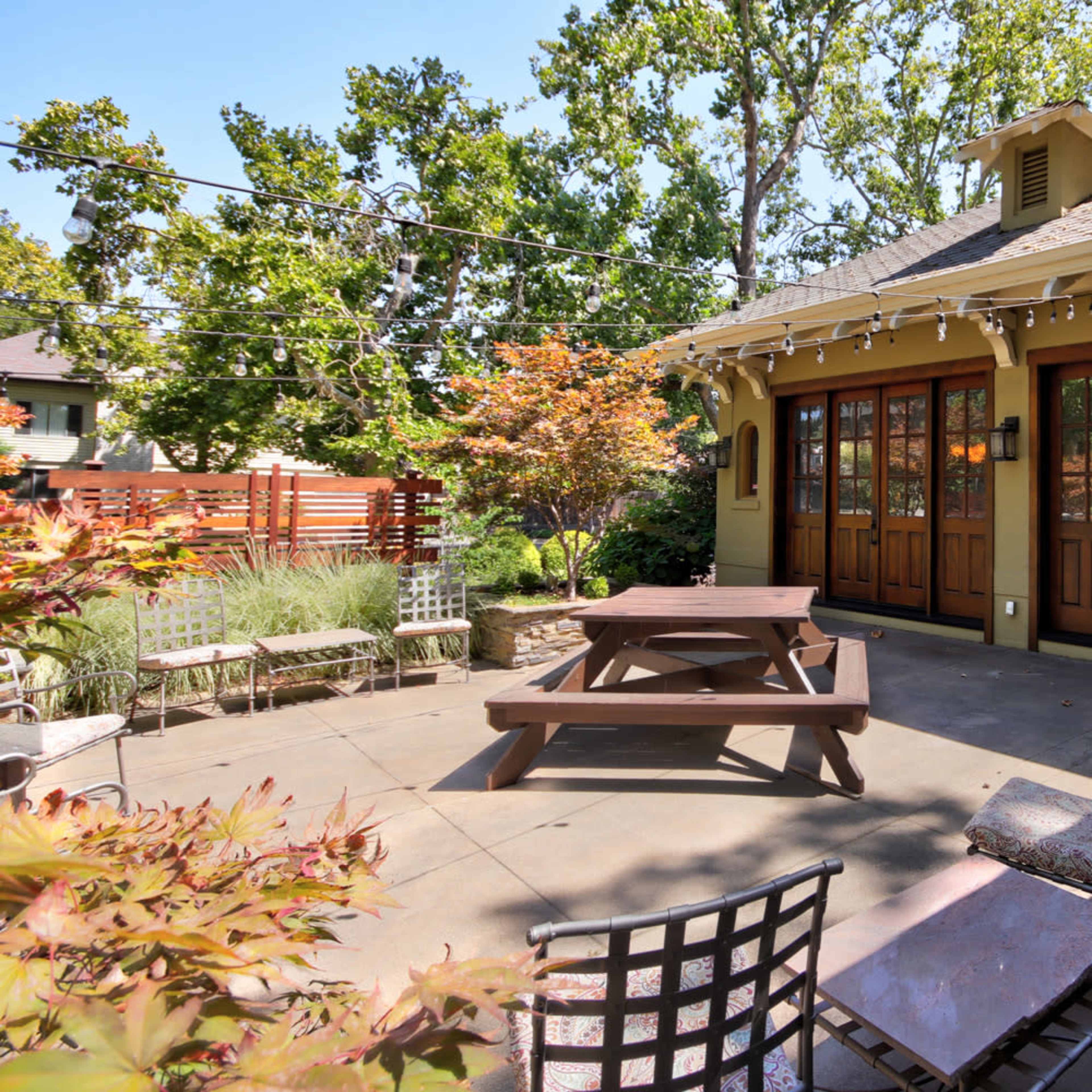 A patio area with a wooden picnic table, several chairs, and landscaped greenery, adjacent to a house with large glass doors.
