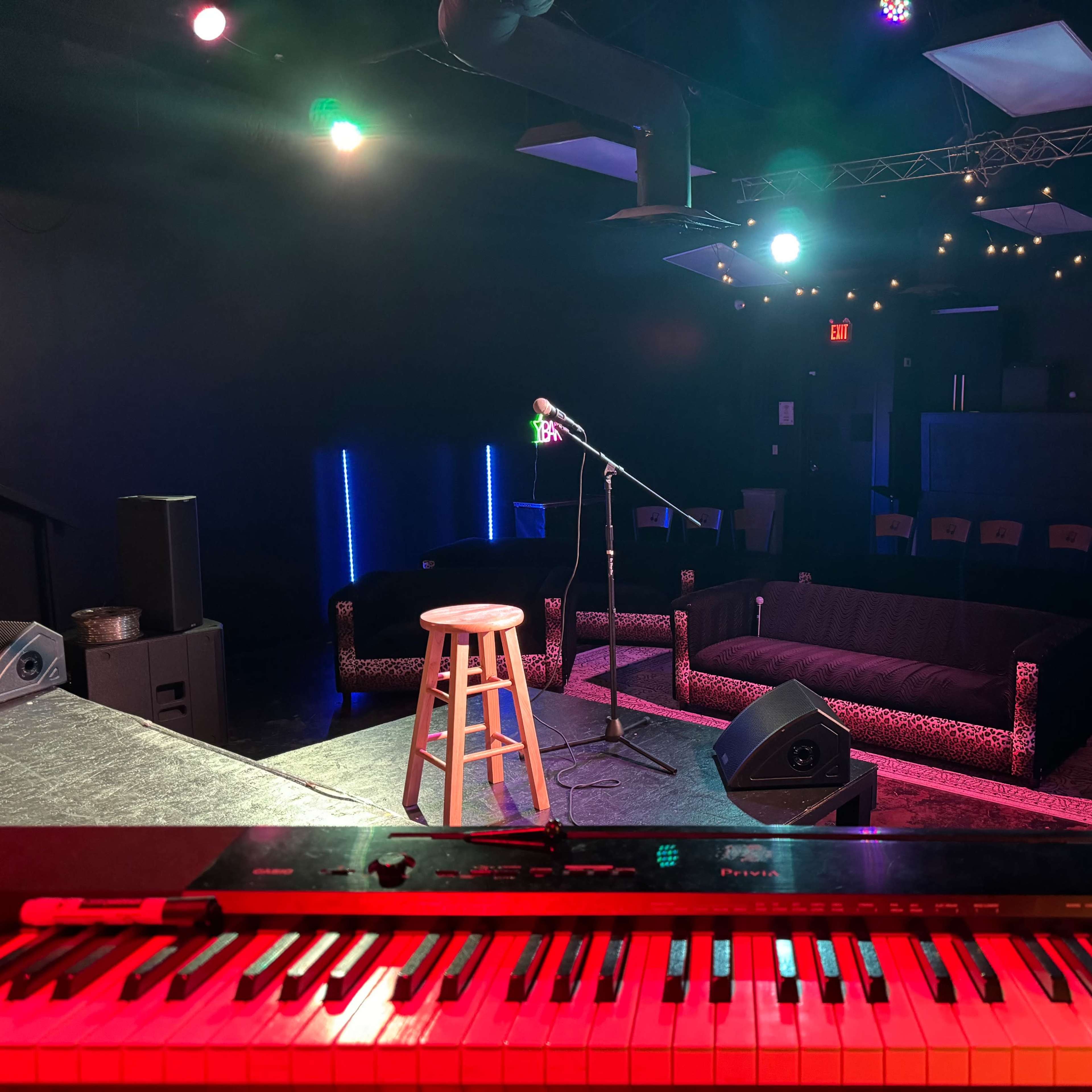 A stage view of a small performance venue with a microphone on a stand, a wooden stool, and a keyboard in the foreground, surrounded by couches and stage lights.