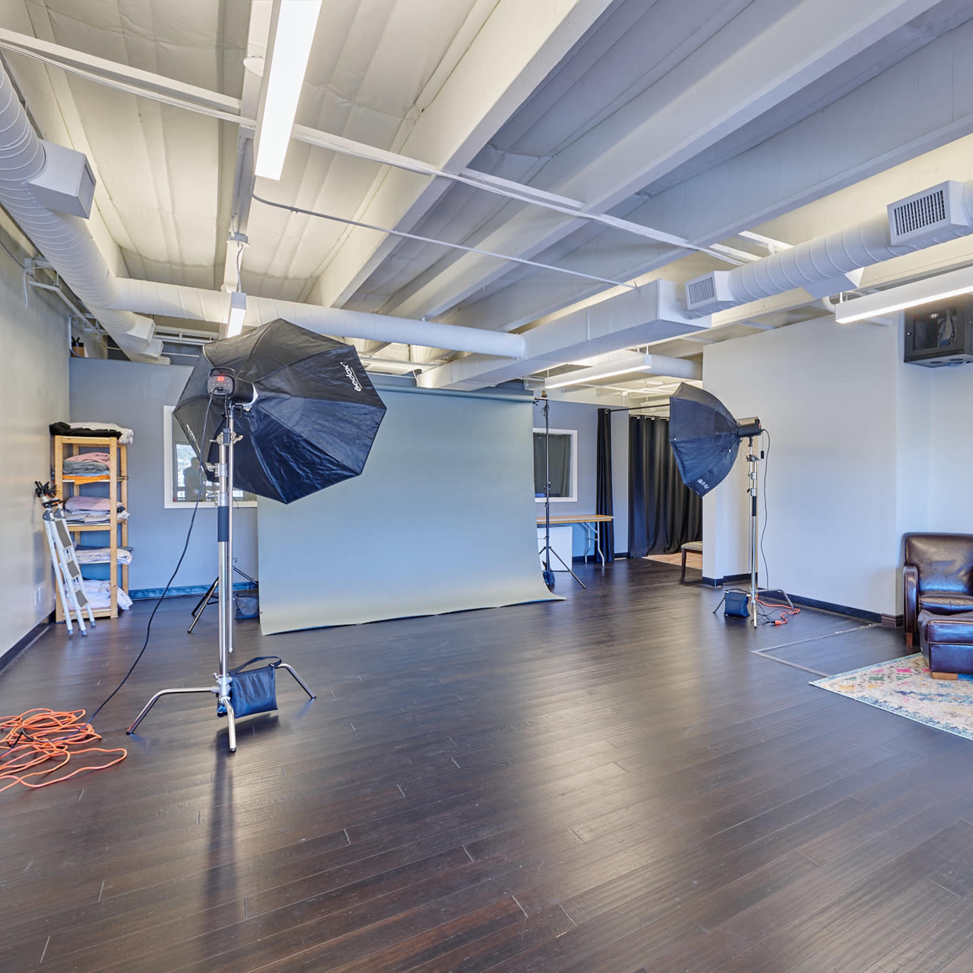 A spacious studio interior equipped with photography lighting, a backdrop, and a seating area with a sofa and armchair.