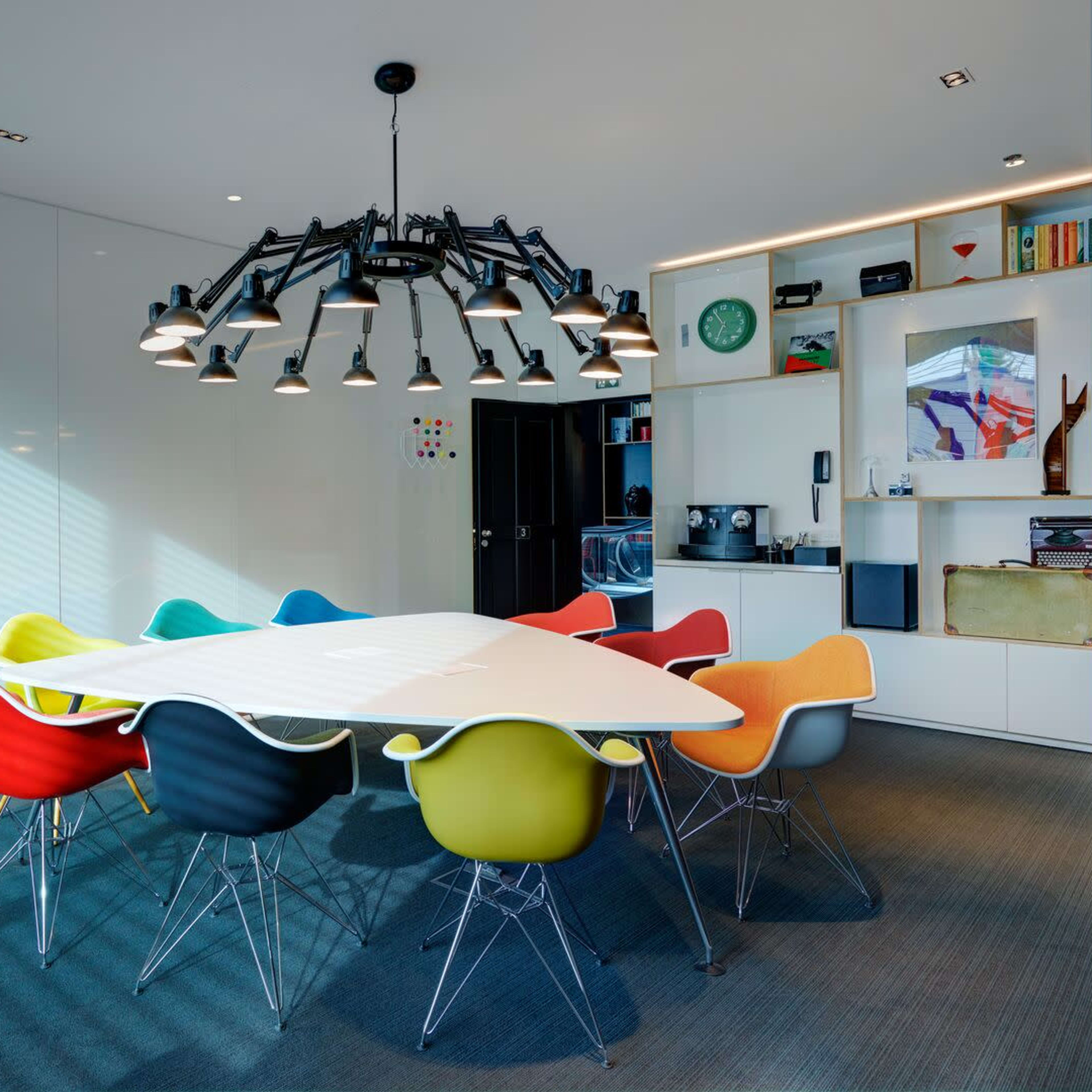 A modern conference room features a large triangular table surrounded by colorful chairs and a striking chandelier above.