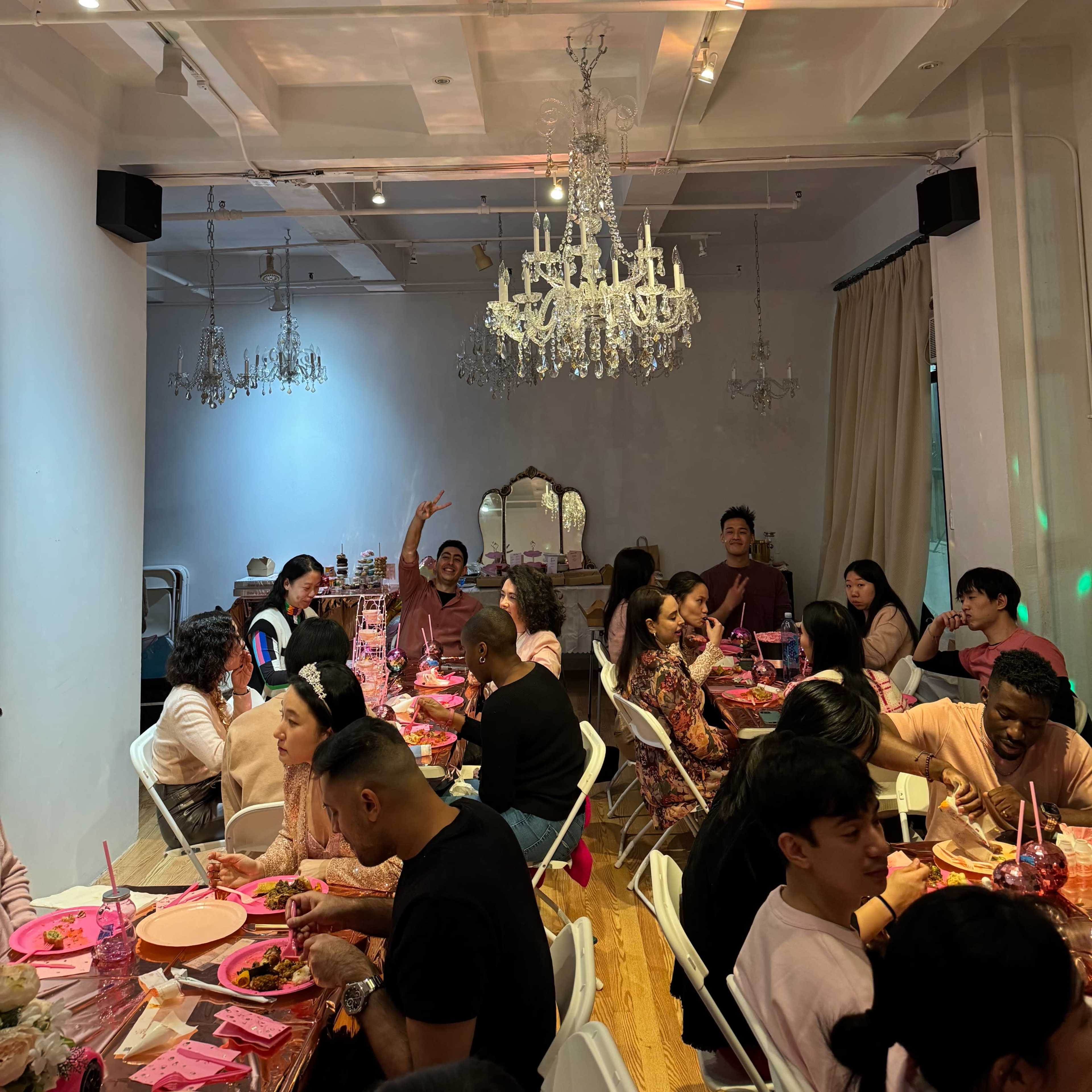 A group of people sits around tables in a brightly lit room, enjoying a meal under a chandelier.