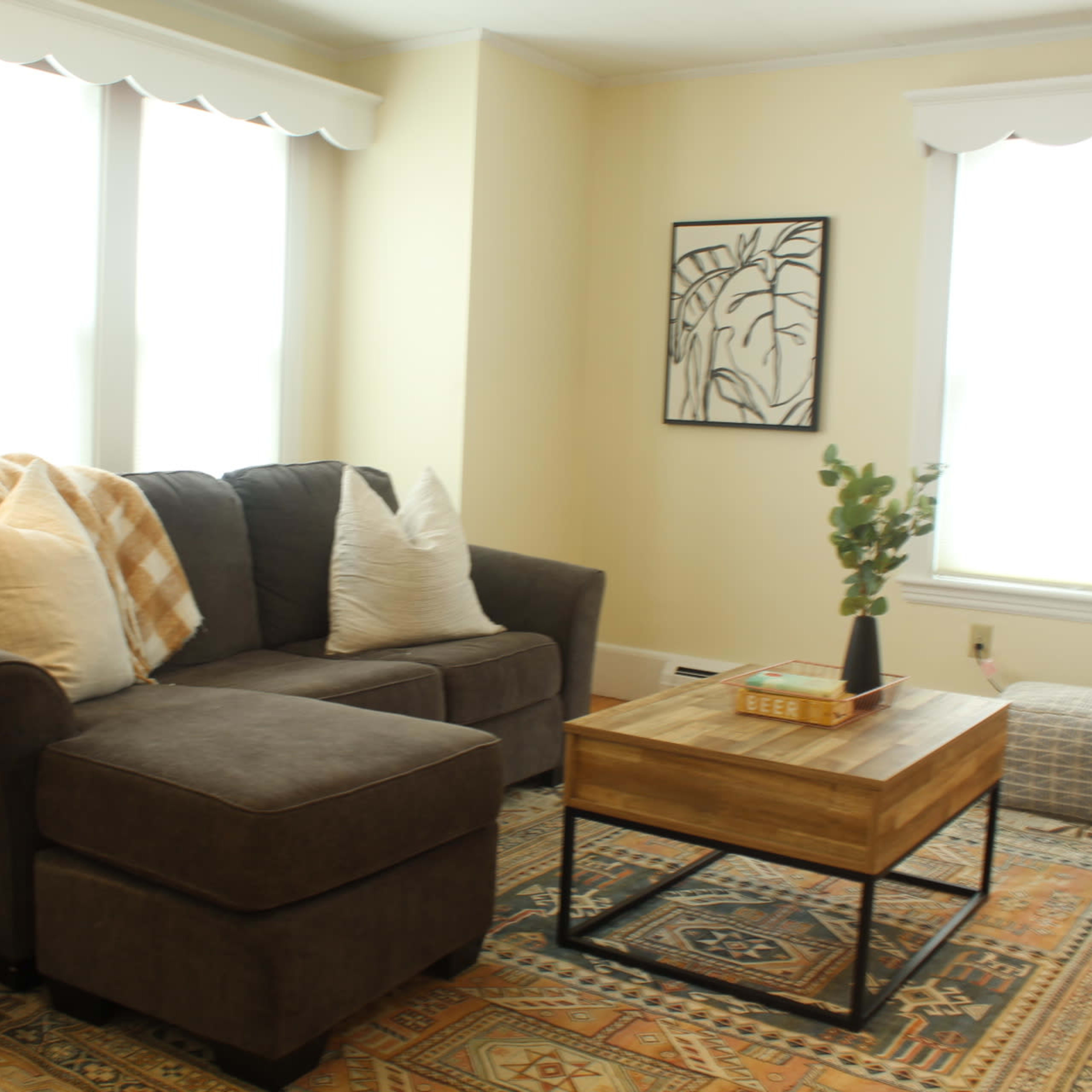 A cozy living room with a gray sectional sofa, a wooden coffee table, and light-colored walls, featuring two large windows and a floor lamp.