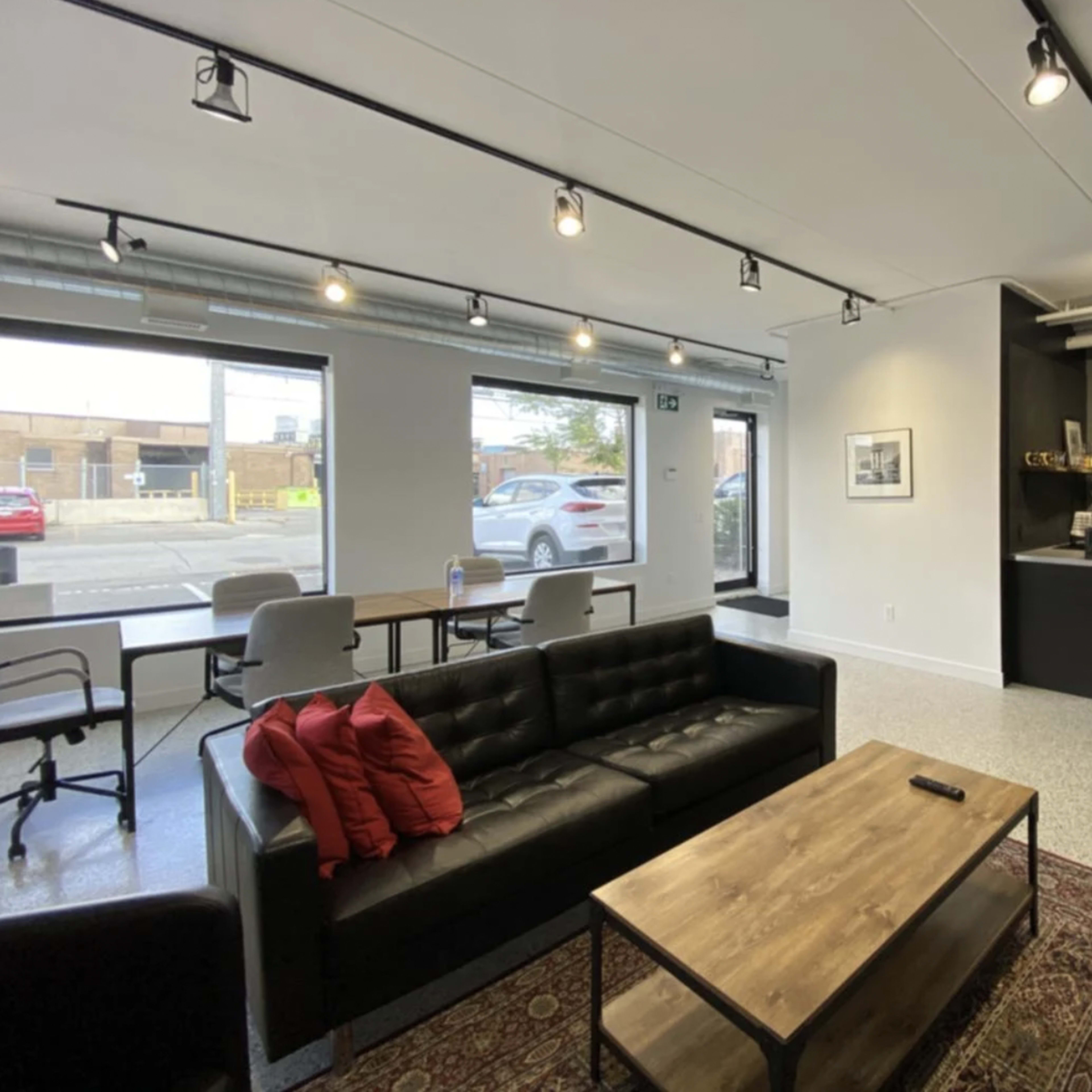 A modern, open lounge area with a black couch, a wooden coffee table, and a kitchenette in the corner, featuring large windows allowing natural light to enter.