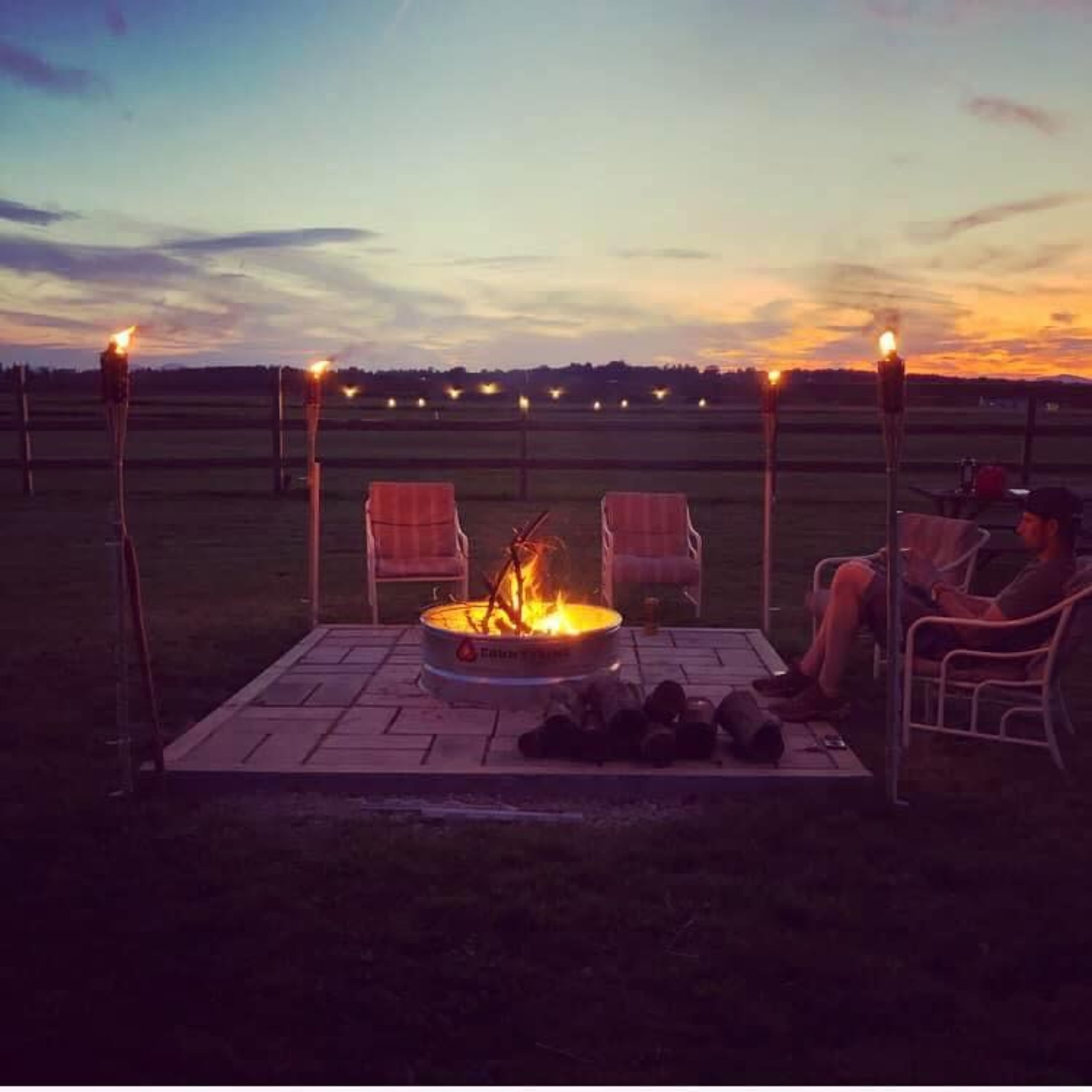 A person sits near a fire pit surrounded by torches and lawn chairs under a colorful sky at dusk.