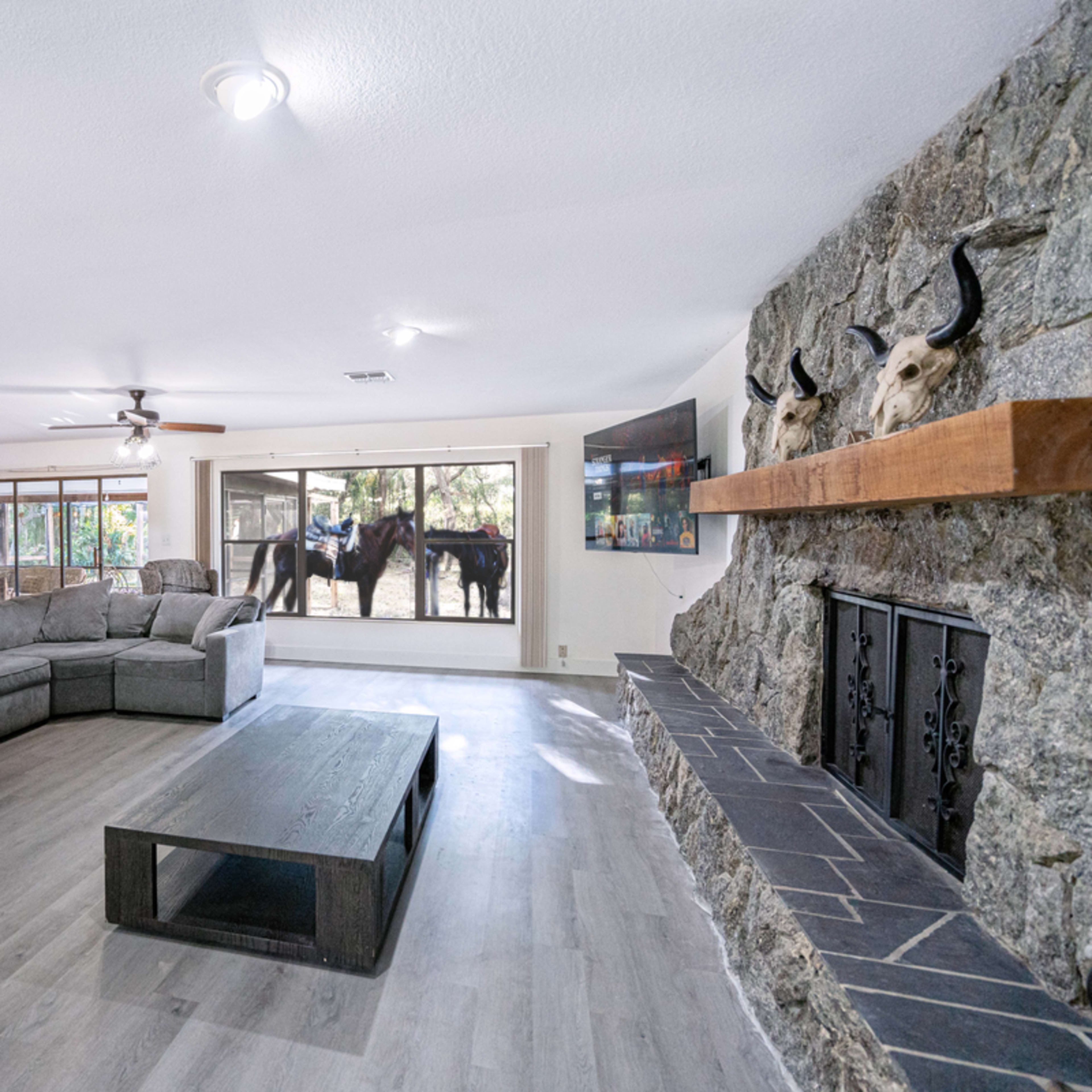 A spacious living room features a large sectional couch and a stone fireplace, with a view of horses outside through large windows.