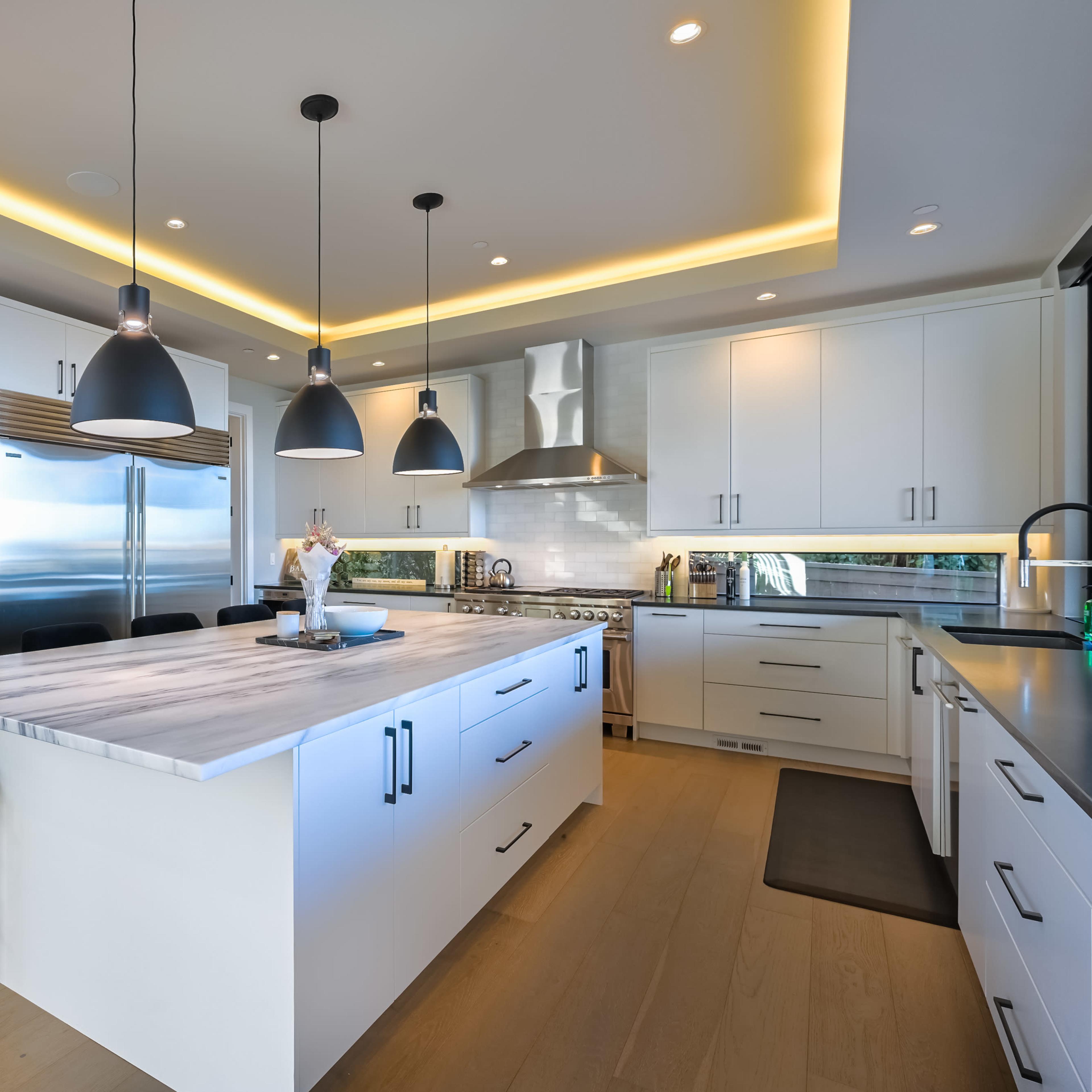 A modern kitchen features sleek white cabinetry, a large marble island, and pendant lighting above, with stainless steel appliances and a window showcasing the outdoors.