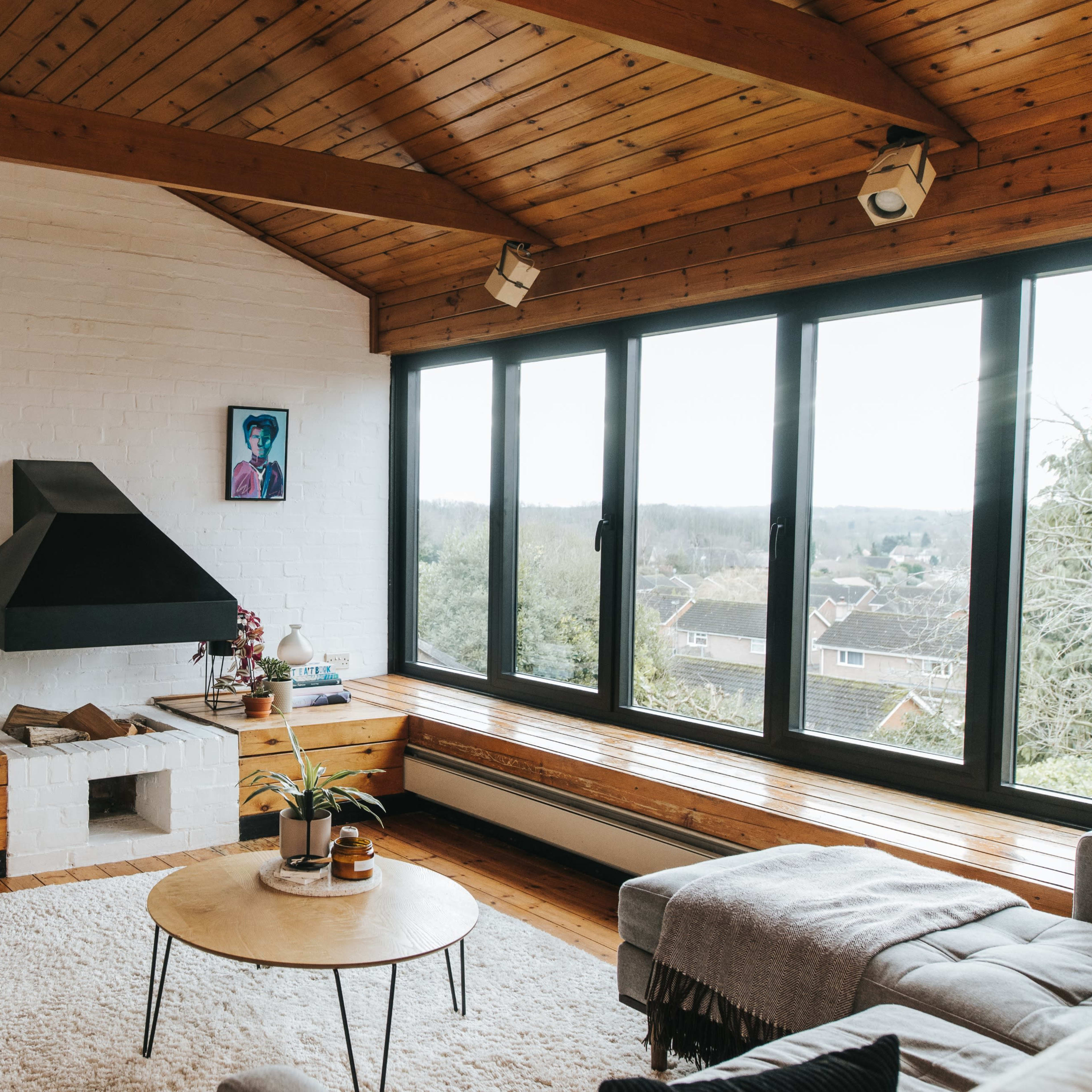 A modern living room features a wooden ceiling, large windows overlooking a hillside, a fireplace, and minimalistic furniture.