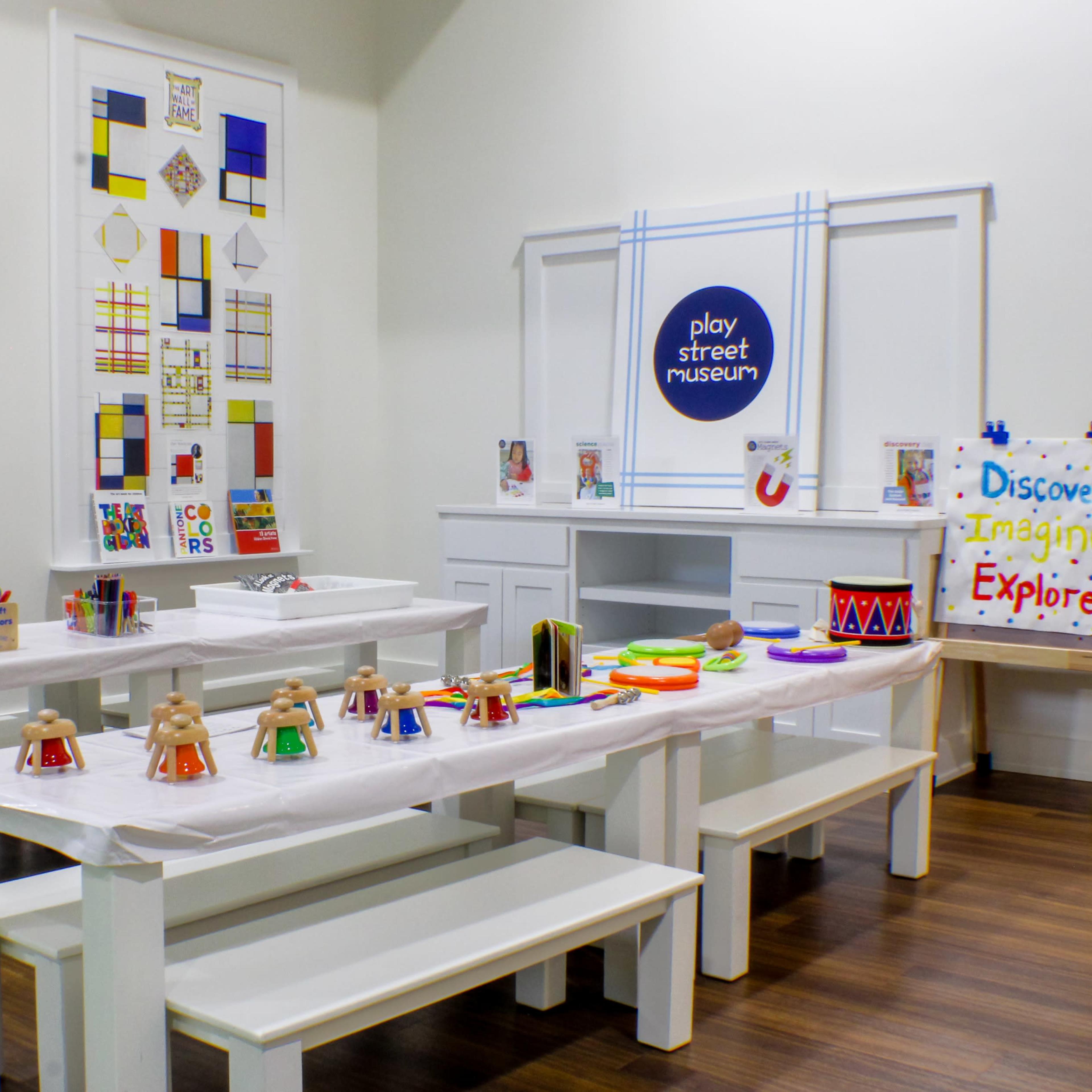 A colorful children's art and activity room with white tables, benches, and various craft supplies arranged on the tables.