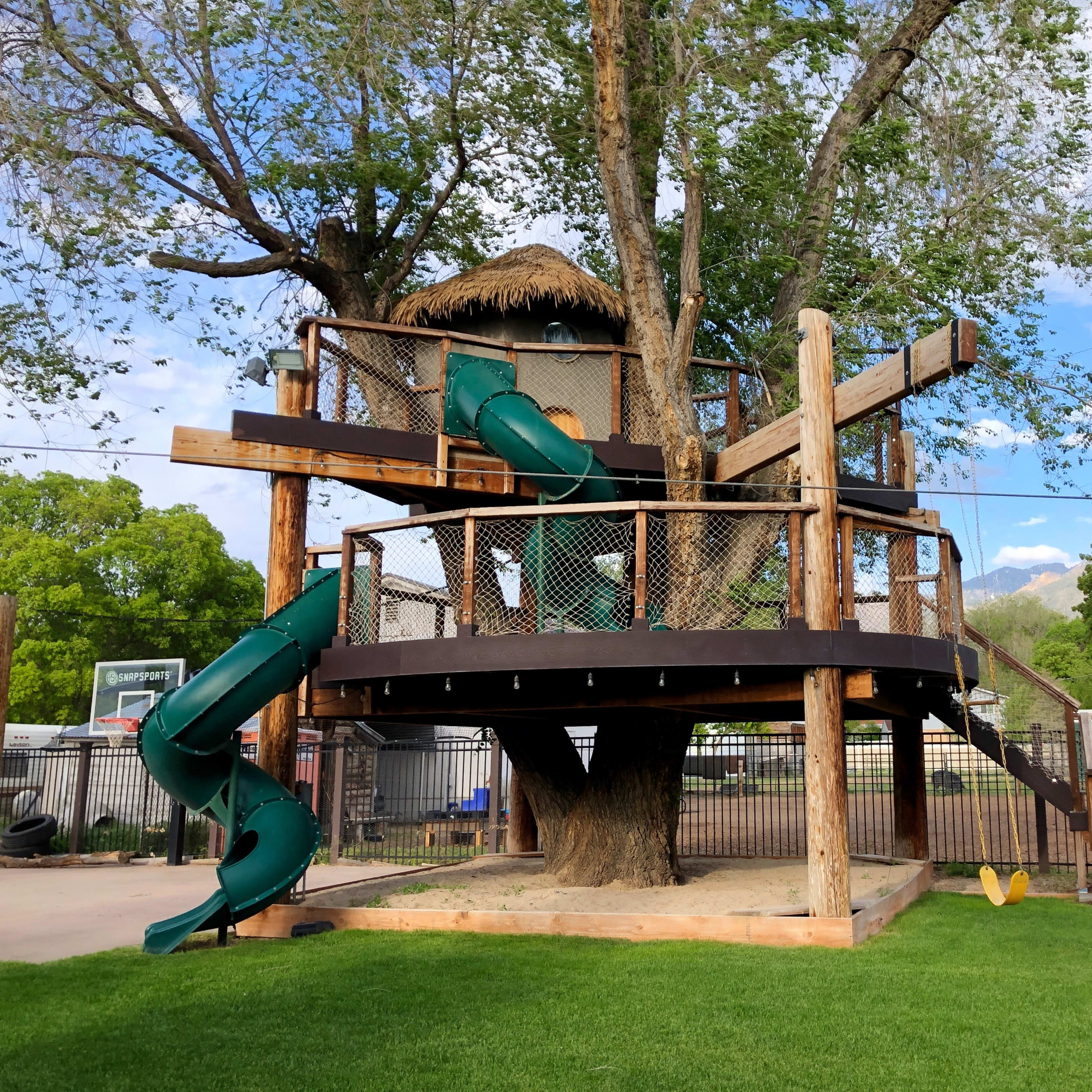 A large wooden treehouse with green slides, situated in a grassy area surrounded by trees and a fence.