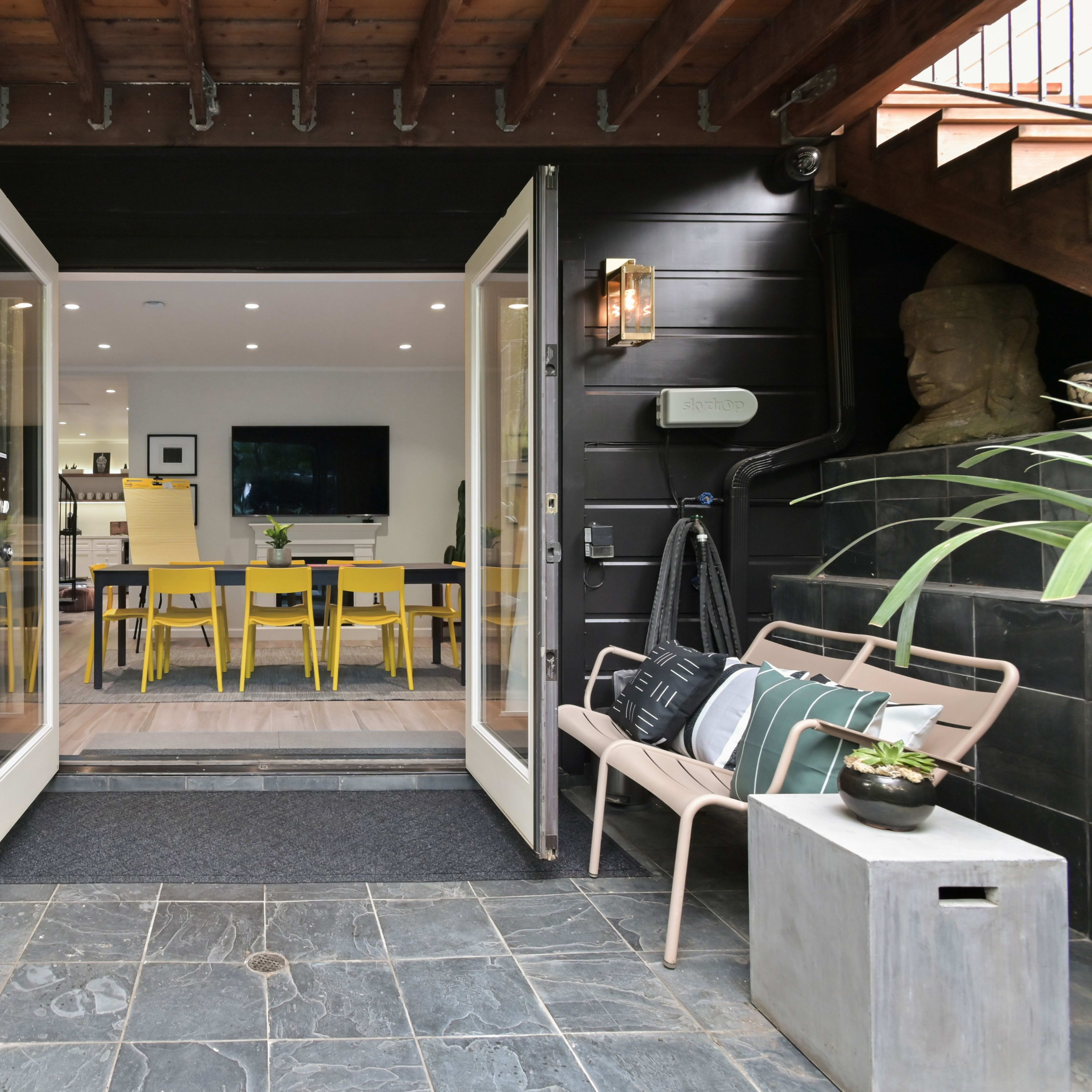 A set of double doors open into a modern dining area with yellow chairs, surrounded by greenery and natural stone features.