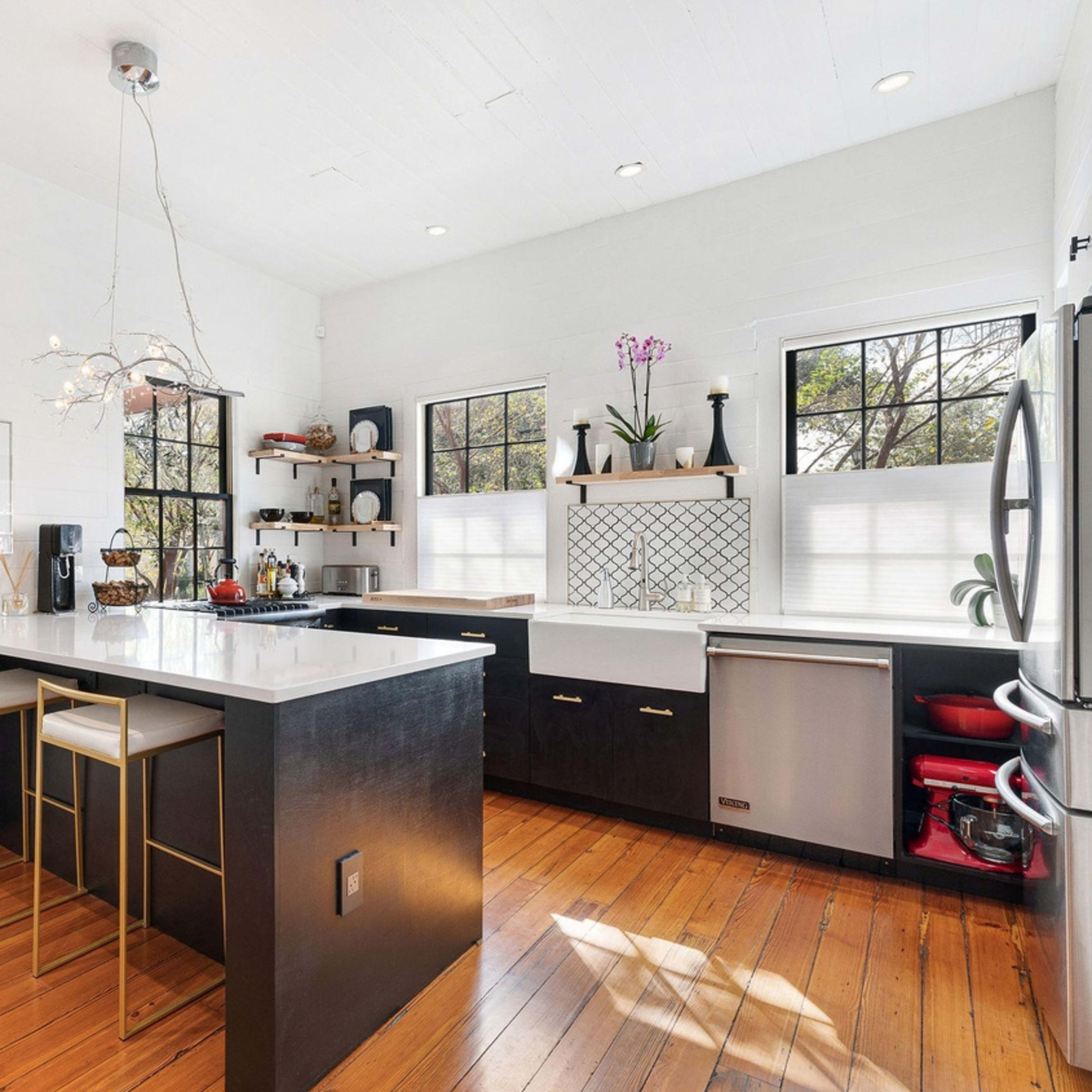 A modern kitchen featuring a large island with bar stools, black cabinets, and wooden flooring, illuminated by natural light through multiple windows.