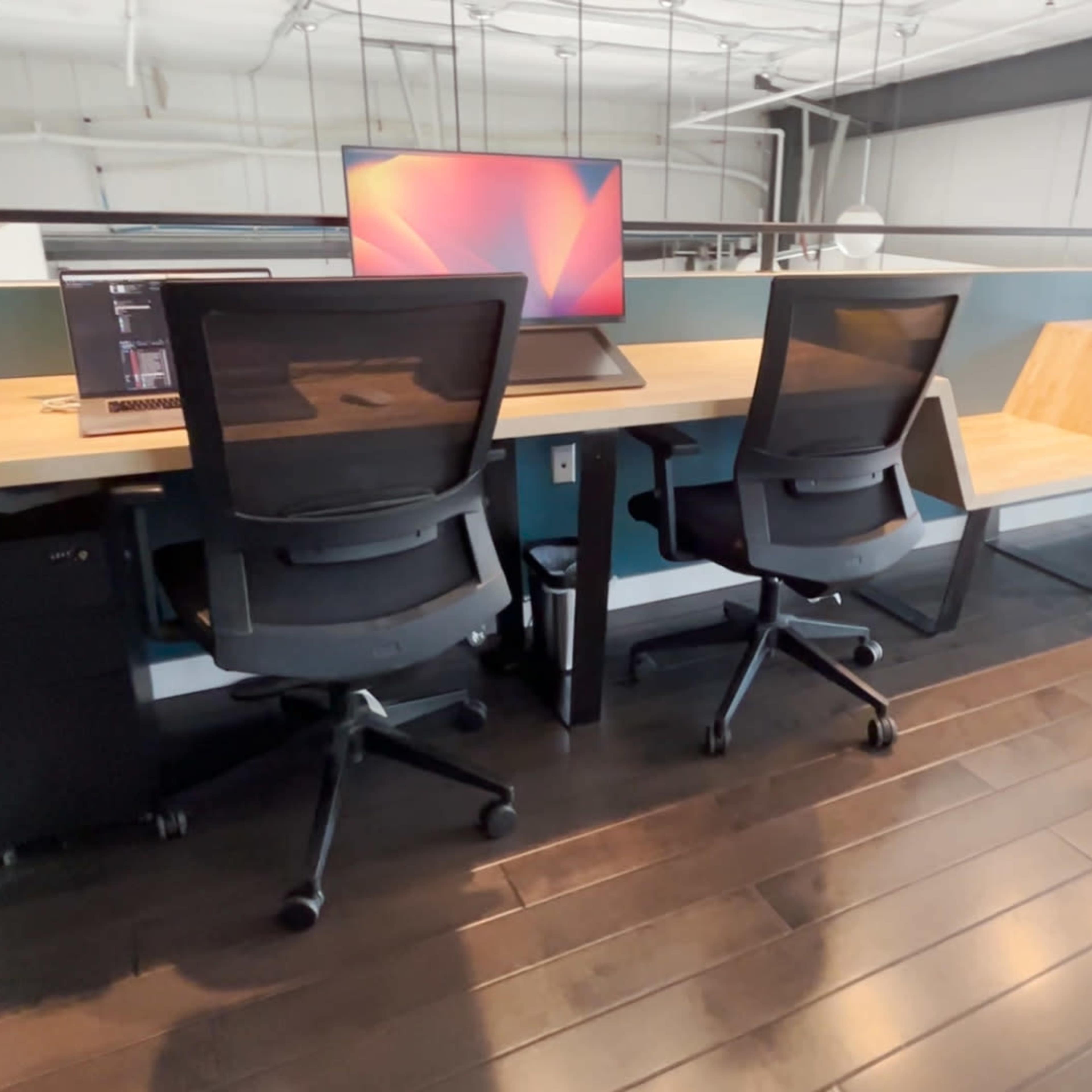 A row of modern office desks equipped with computer monitors and ergonomic chairs is set against a teal wall in a spacious workspace.