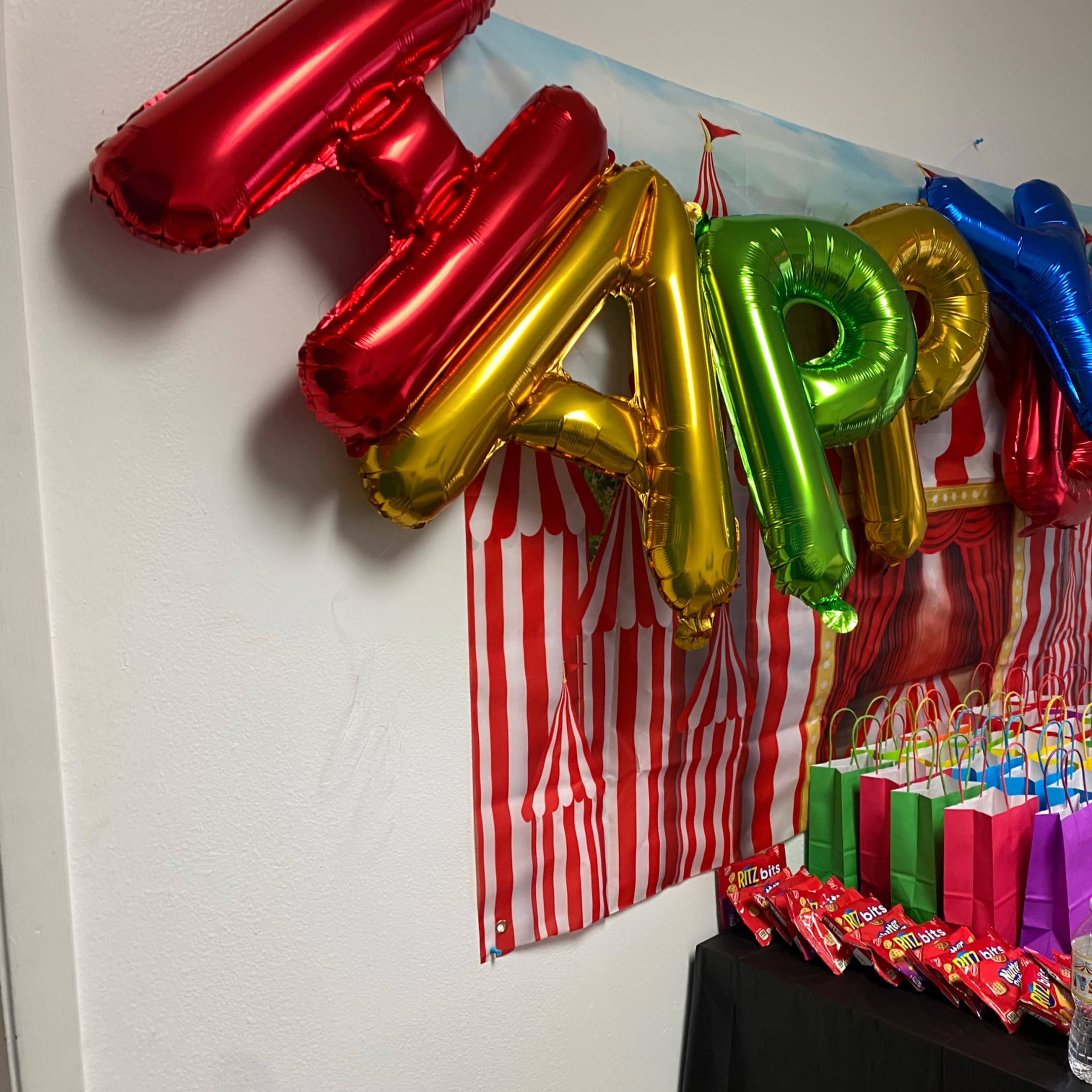 A table displays colorful balloons spelling "HAPPY" alongside a variety of gift bags and candy.