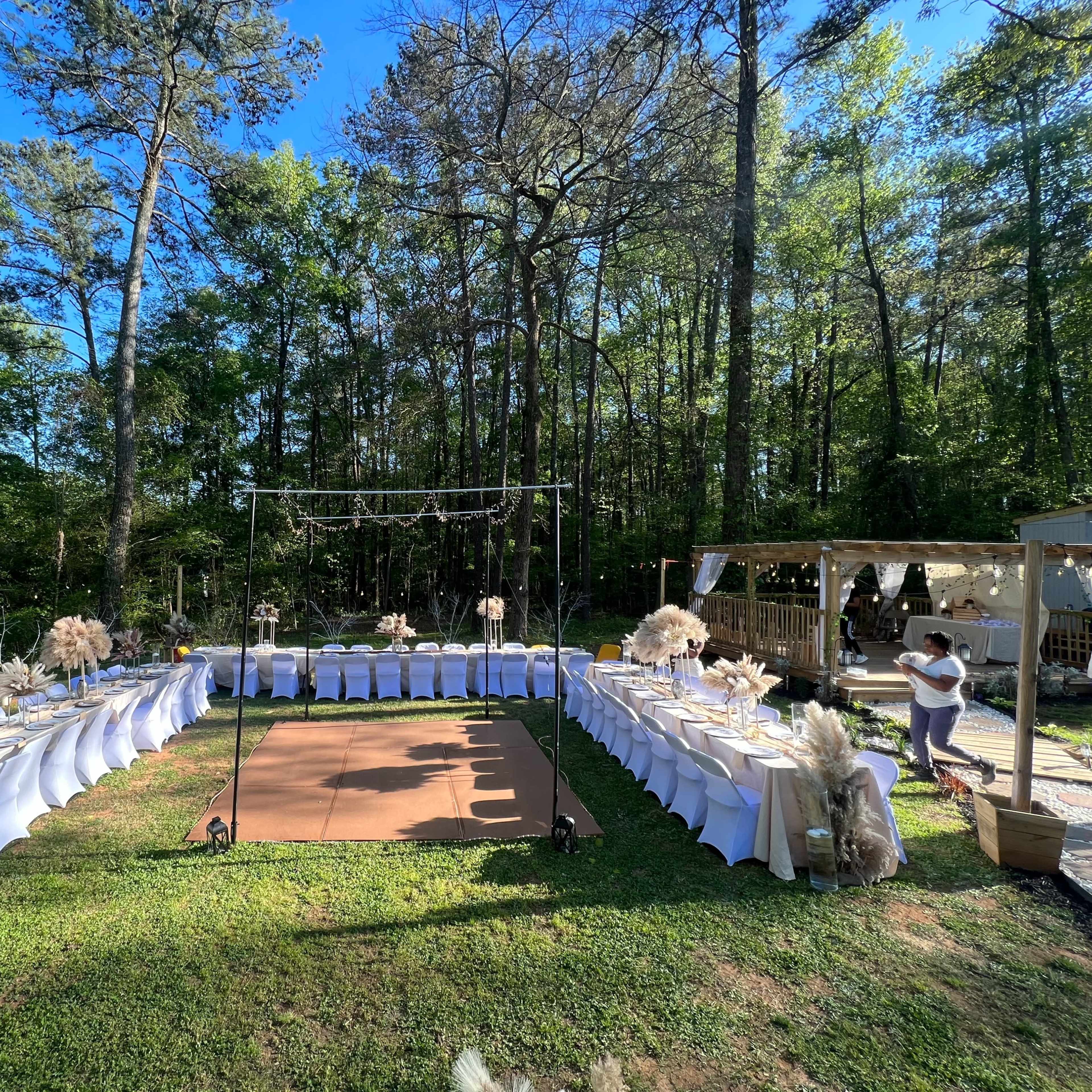 A rectangular outdoor event setup features tables arranged in a curve, surrounded by trees, with a dance floor and a decorated arch in the center.