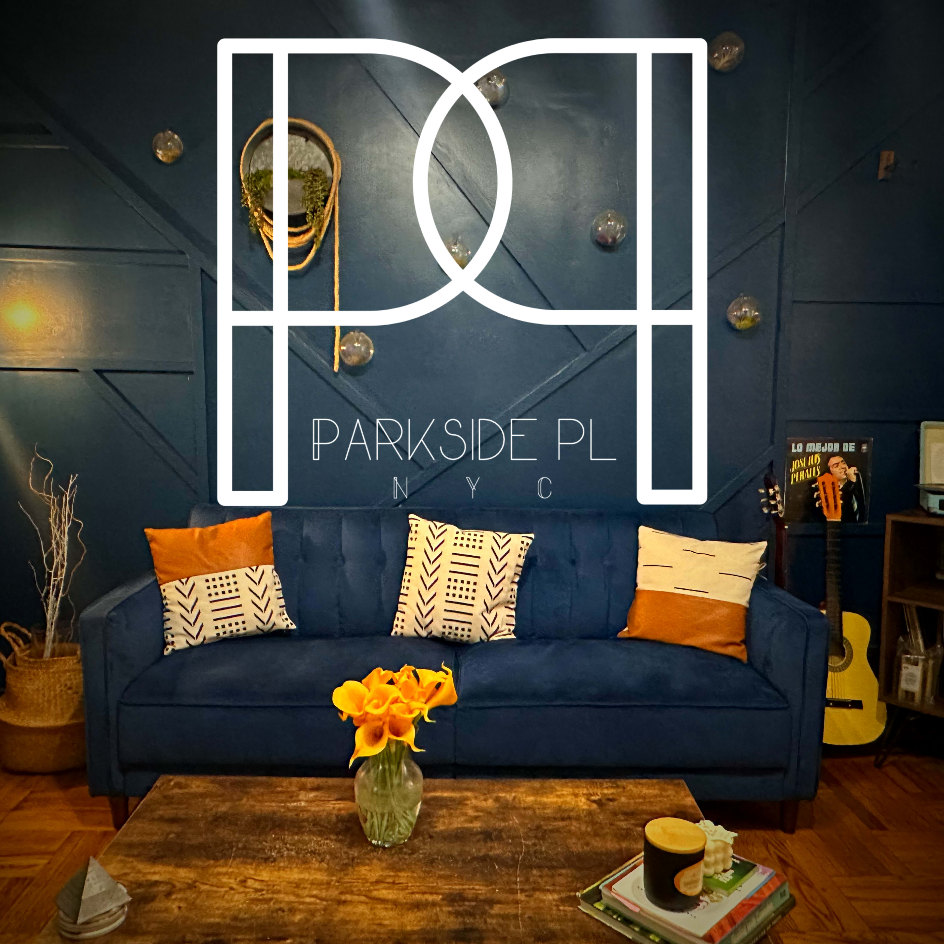 A cozy living room with a blue couch adorned with orange and patterned cushions, a wooden coffee table with a vase of yellow flowers, and a decorative wall featuring a mix of textures and lighting.
