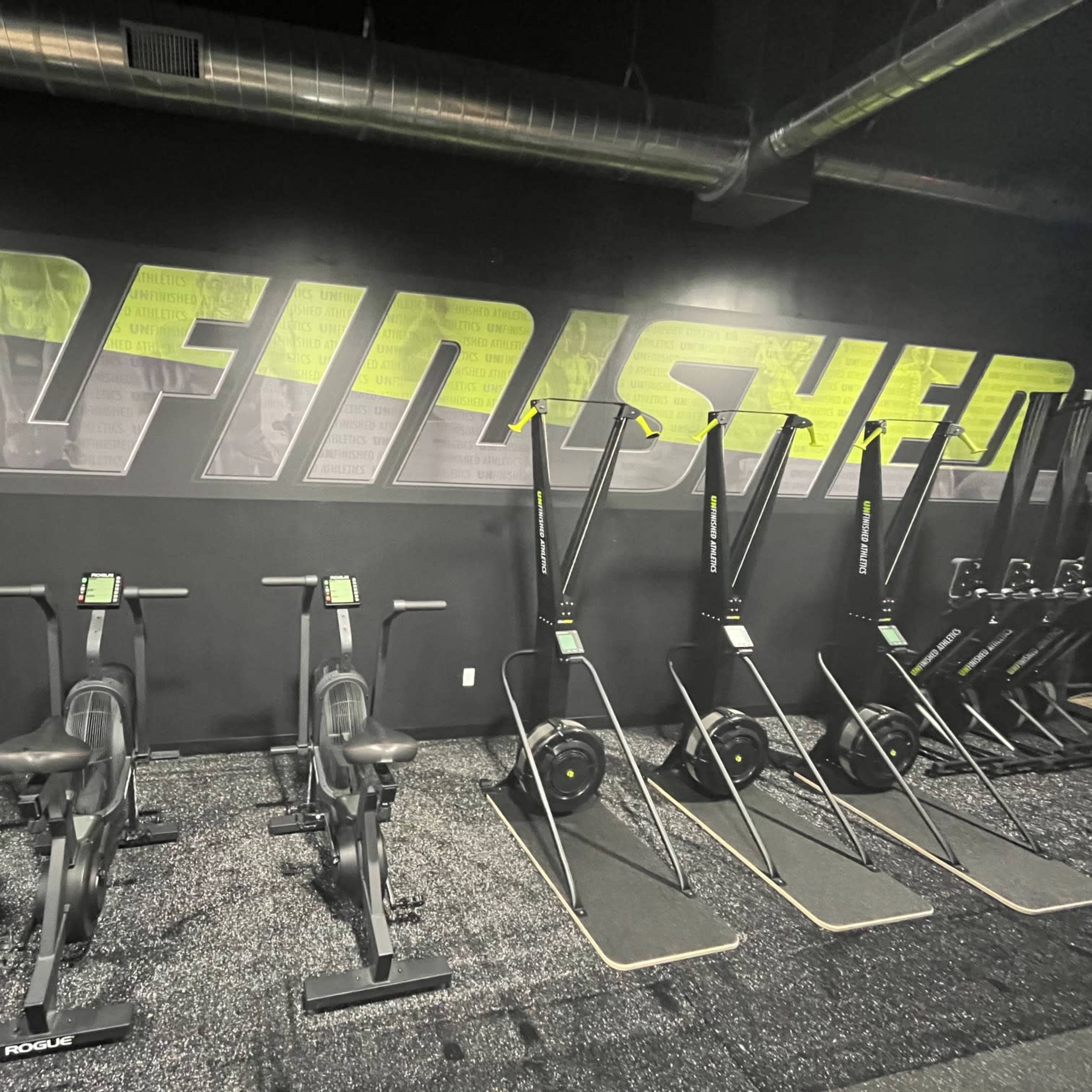 A gym interior featuring rowing machines and exercise equipment lined up against a dark wall with the word "UNFINISHED" displayed in bold letters.