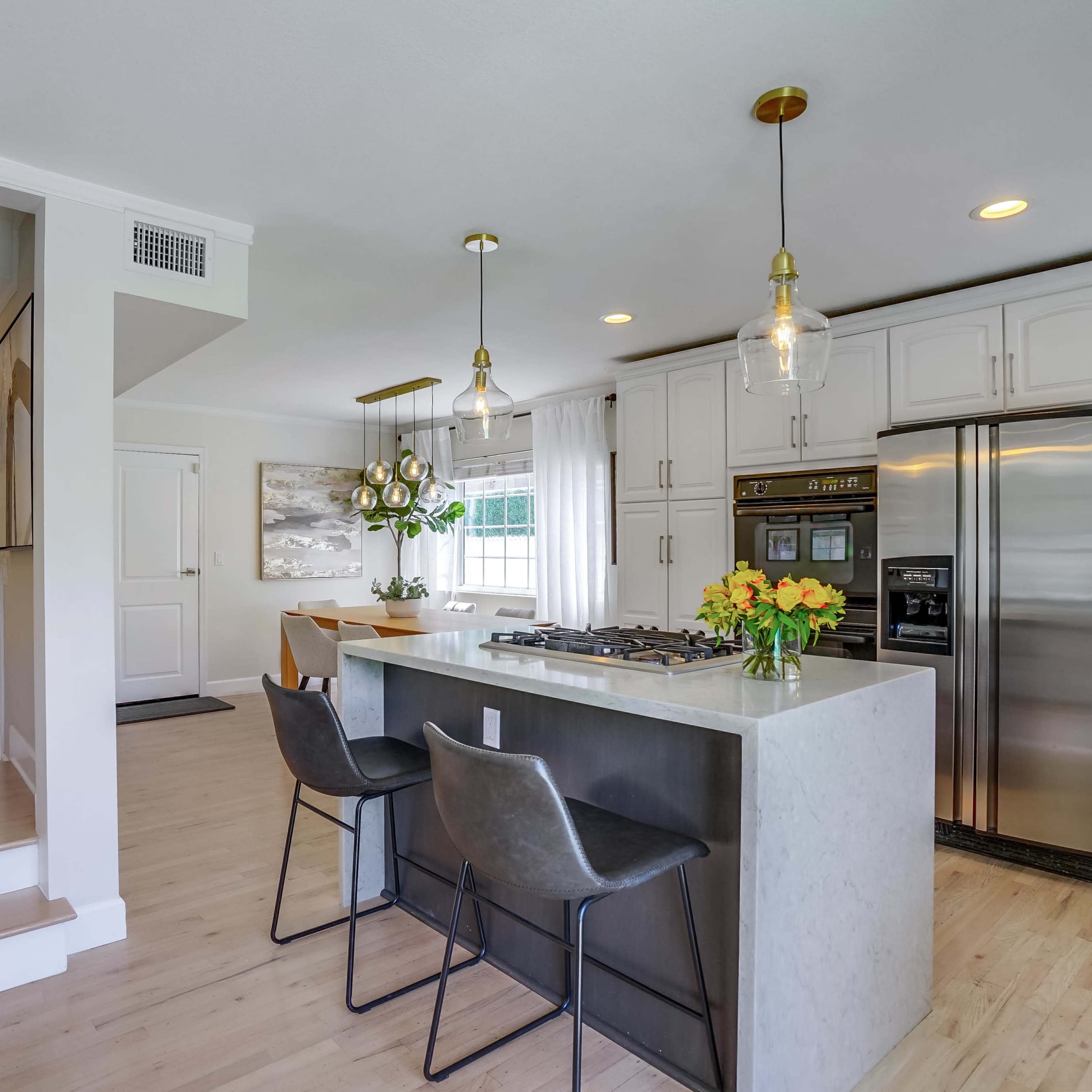 A modern kitchen features a central island with bar stools, stainless steel appliances, and bright natural light coming through a nearby window.