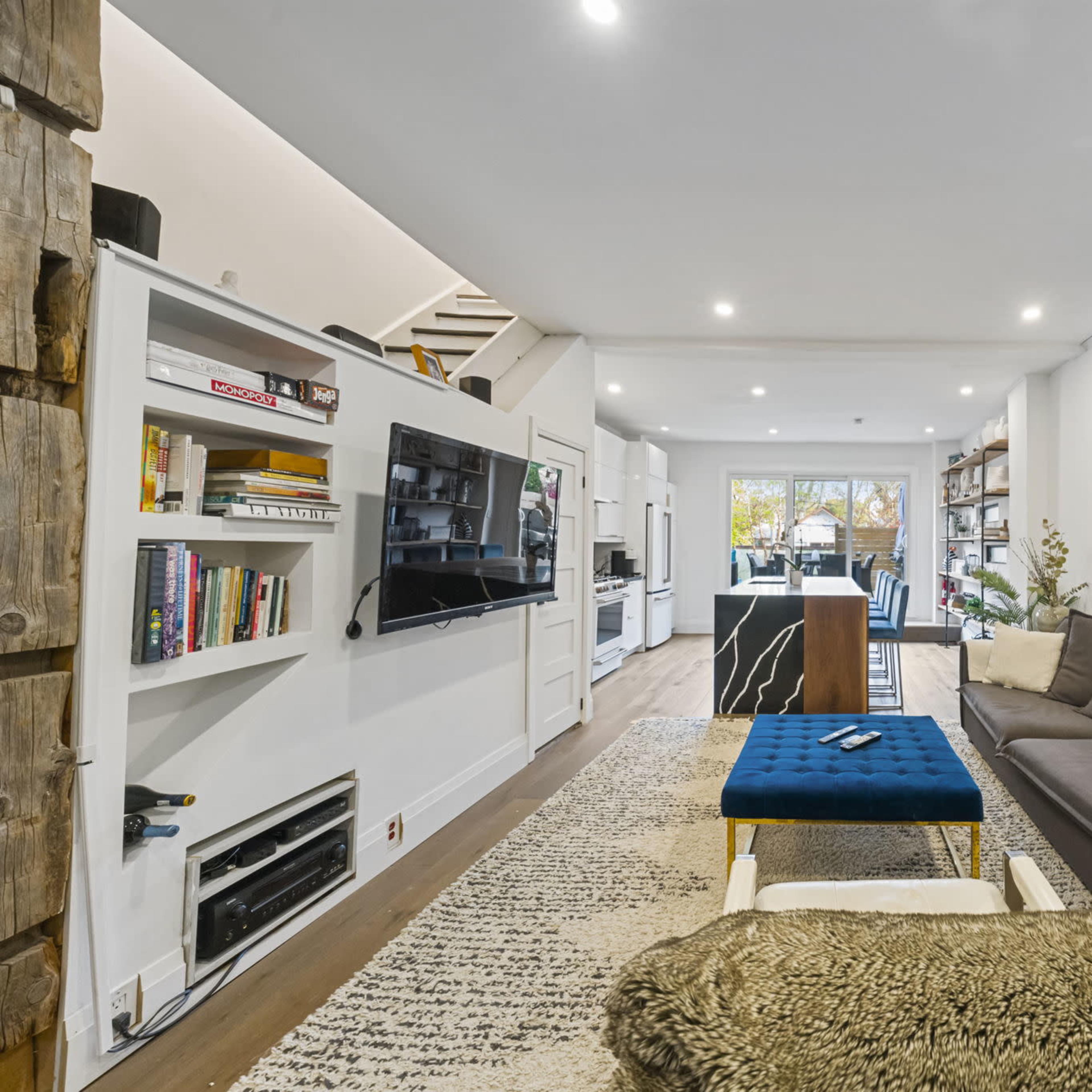 A modern living room with a sofa, a coffee table, shelves filled with books, and a view toward a dining area in the background.