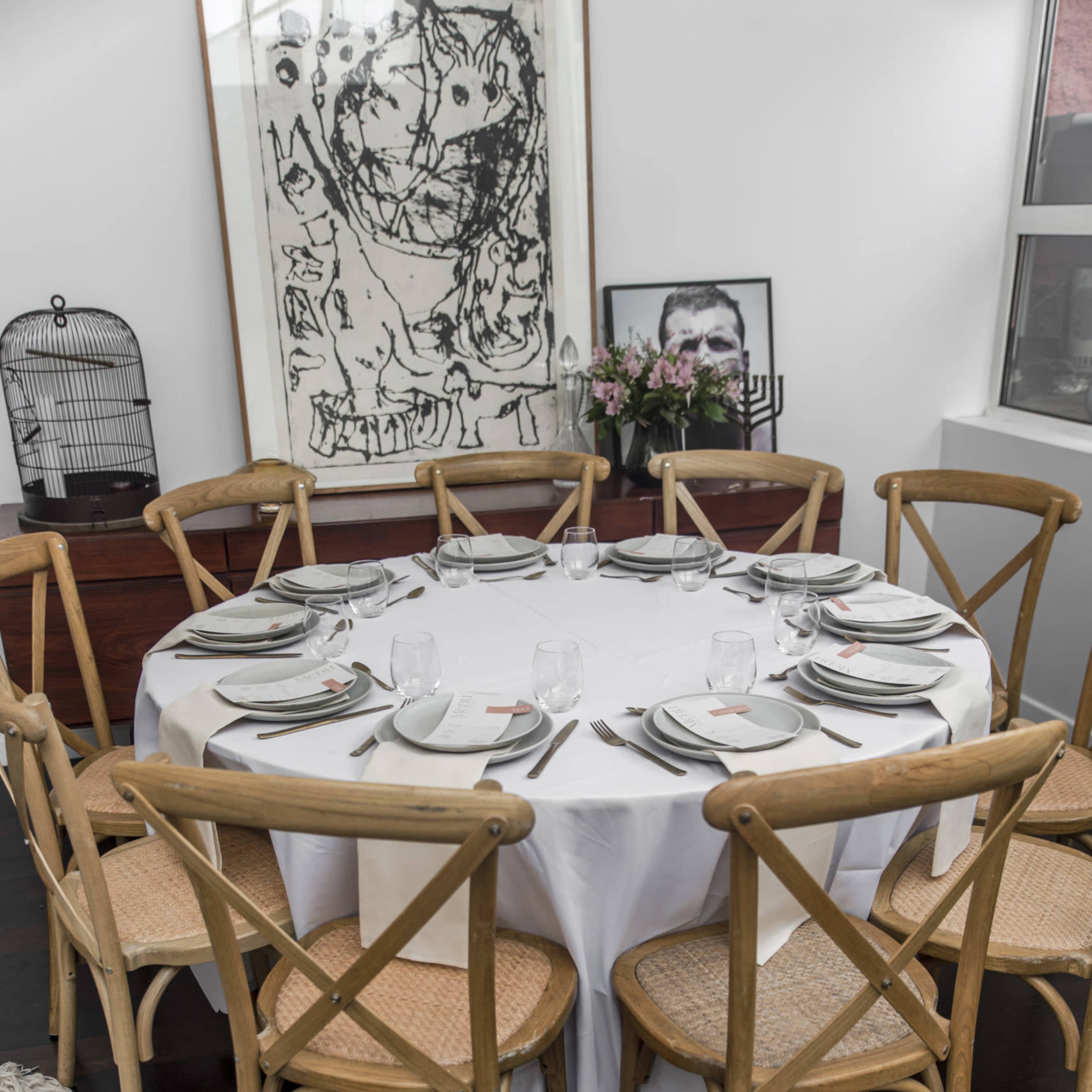 A round dining table is set with plates and glasses, surrounded by wooden chairs, against a backdrop of artwork and a birdcage.