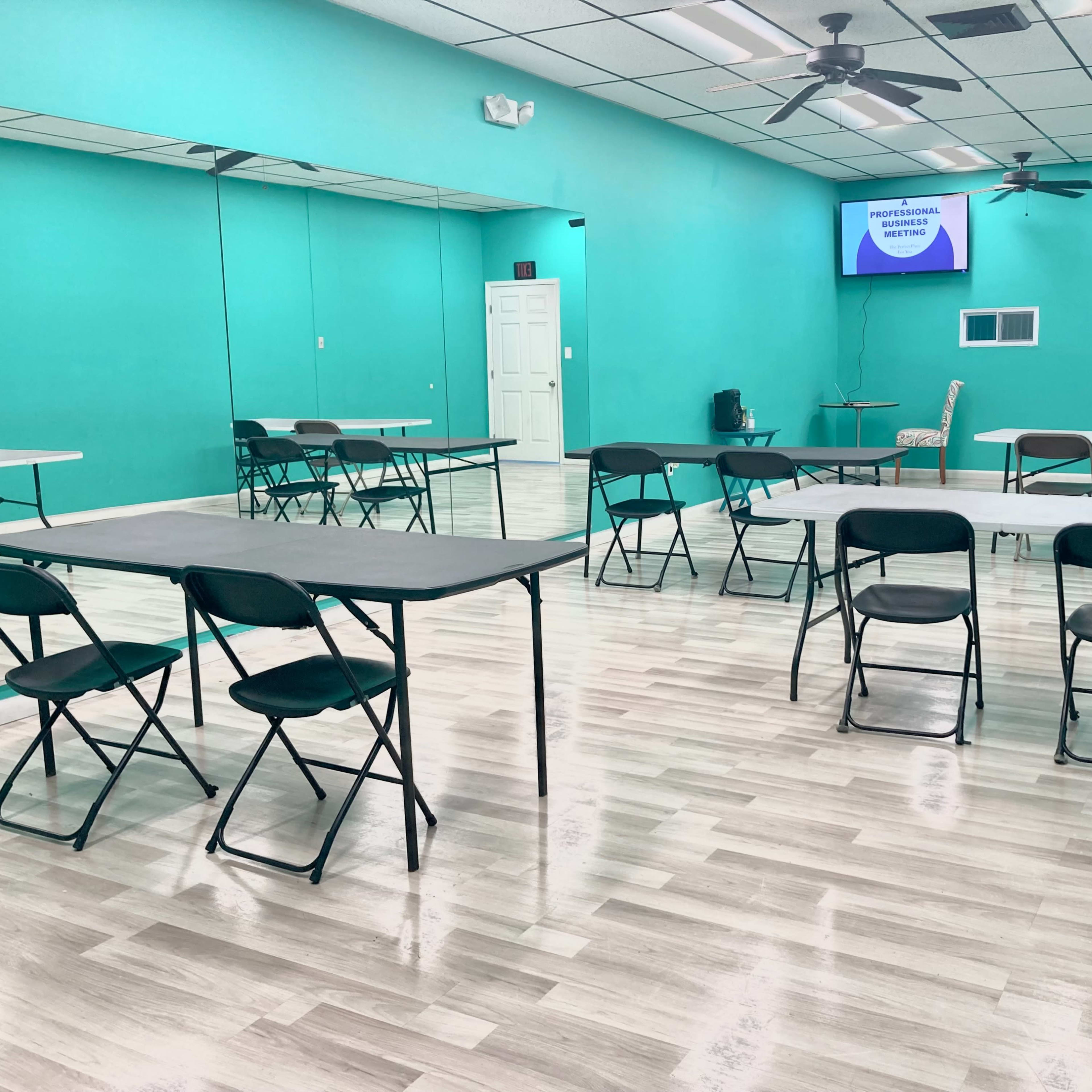 A brightly colored room with teal walls, featuring several folding tables and chairs arranged throughout, and a large mirror on one side.