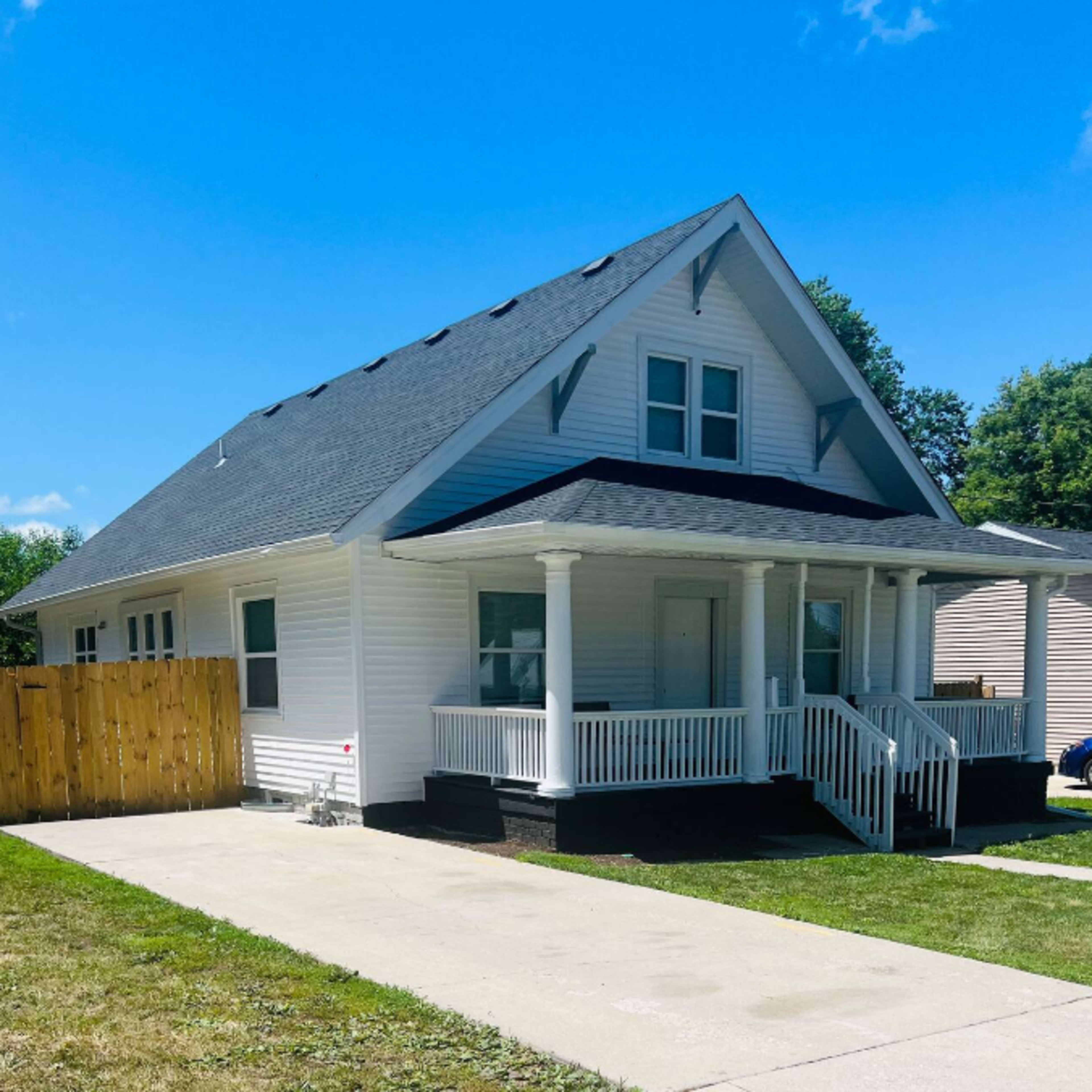 A single-story house with a gabled roof, white siding, and a front porch is situated next to a wooden fence.