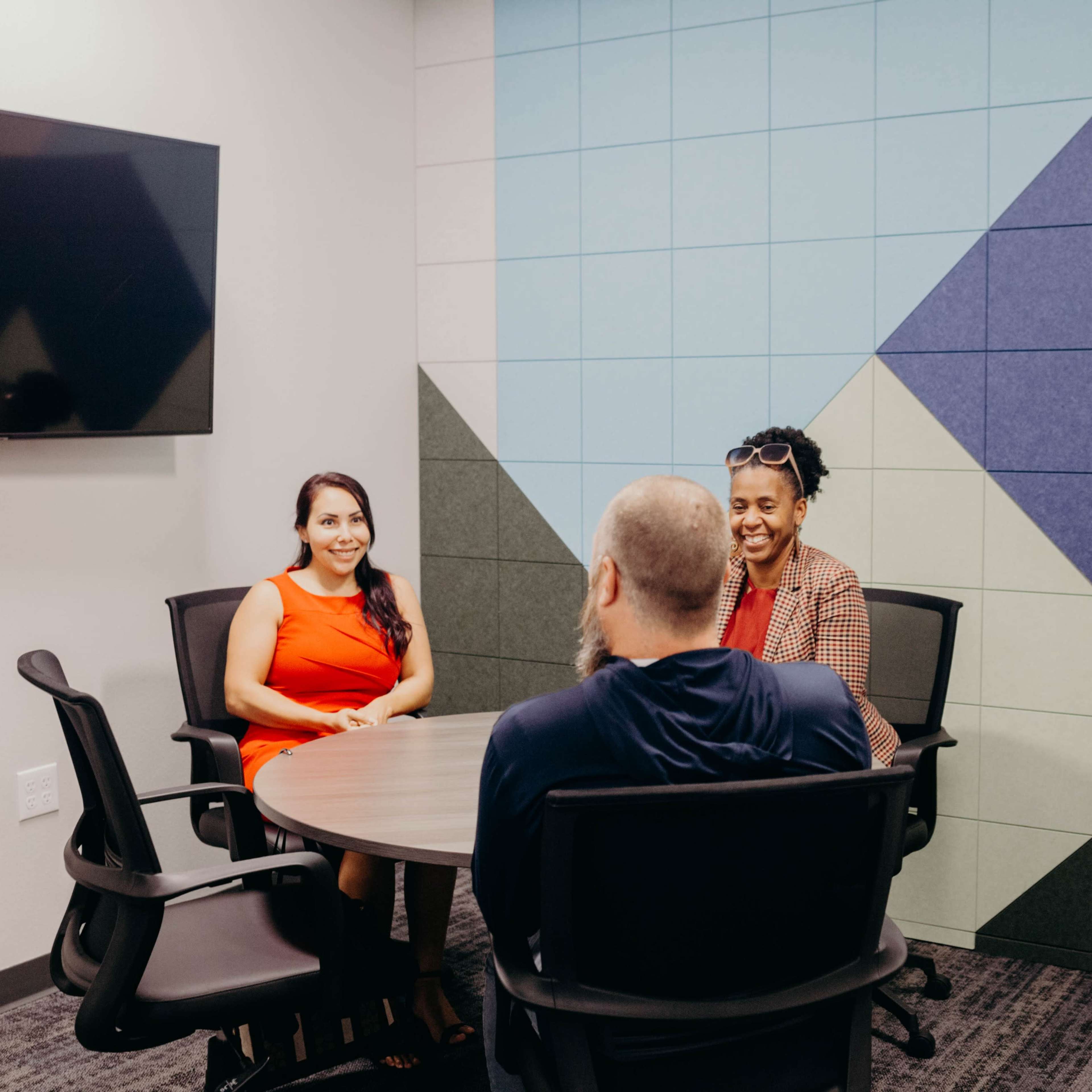 Three people are seated around a circular table in a small meeting room with a colorful wall.