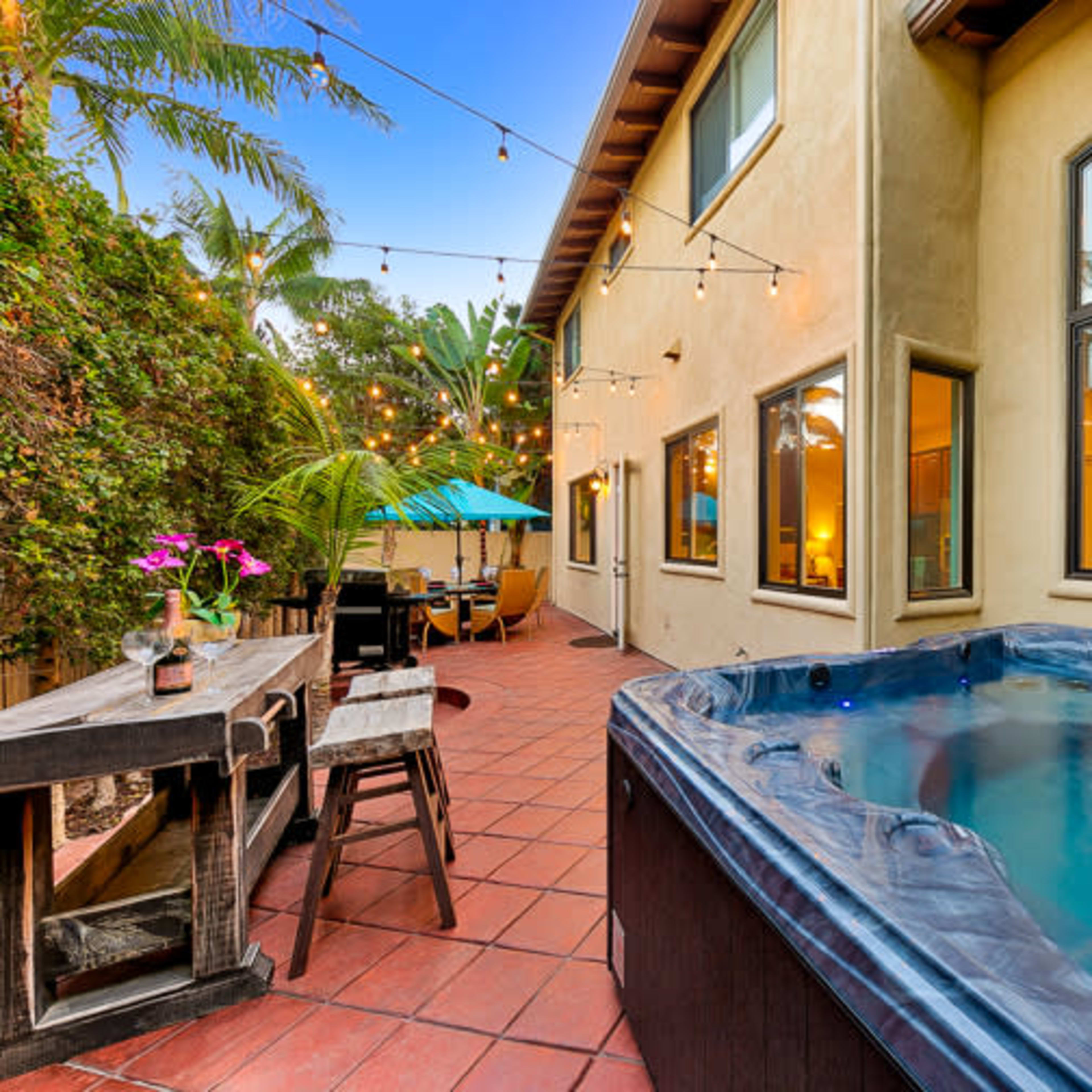 A hot tub is positioned next to a patio area with string lights and outdoor furniture adjacent to a house surrounded by palm trees.