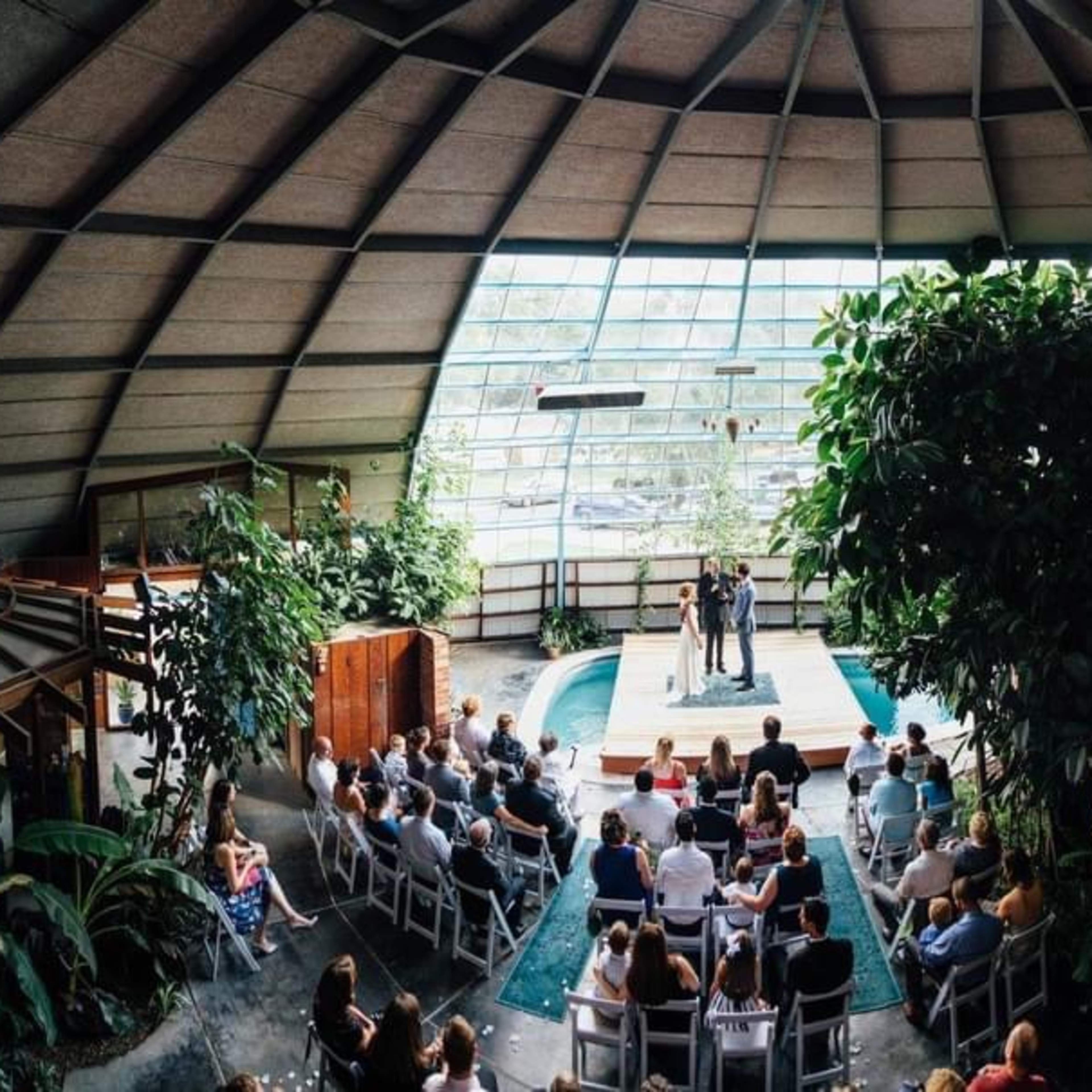 A wedding ceremony takes place indoors under a large, arched glass structure surrounded by tropical plants and a small pool.