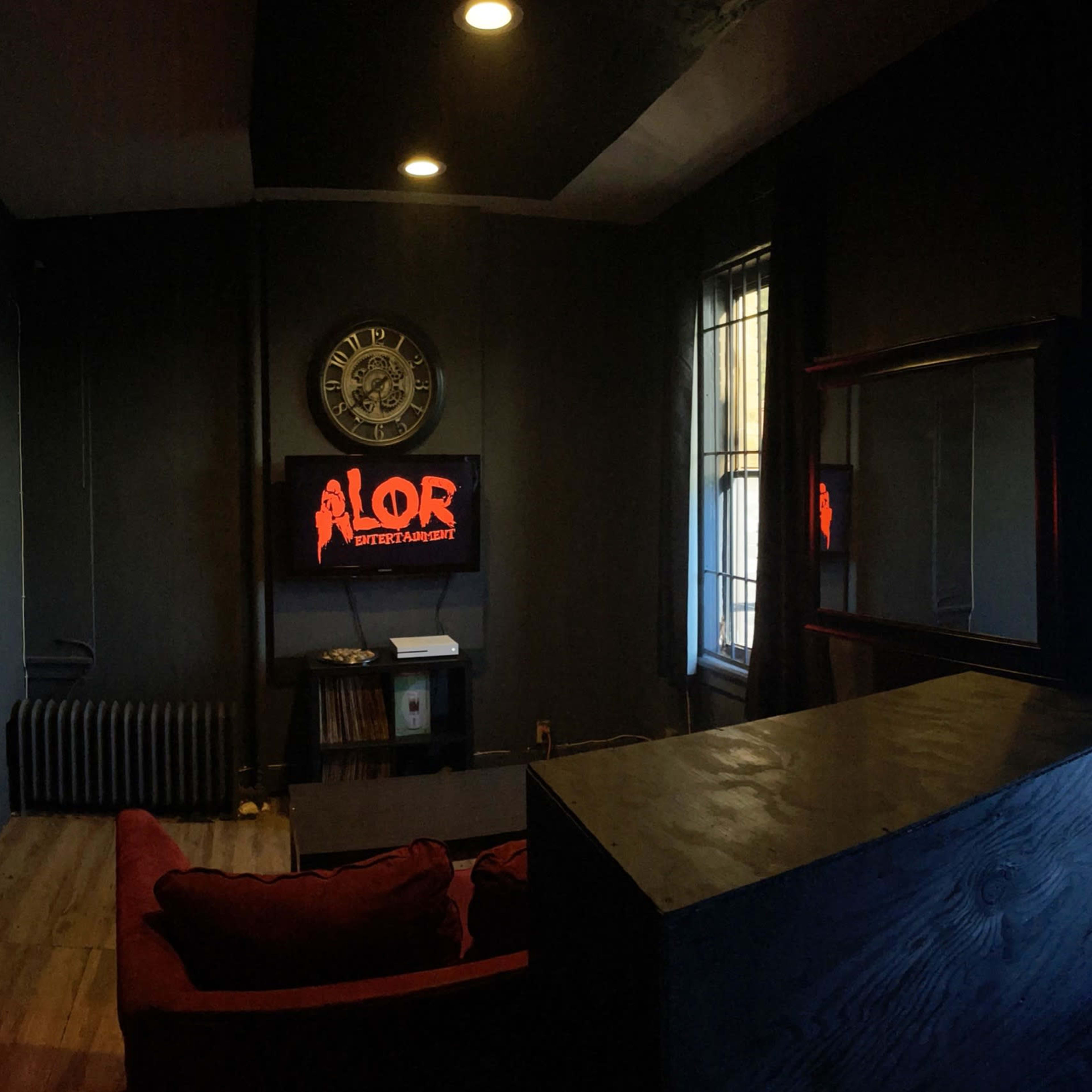 A dimly lit room with dark walls, featuring a red neon sign reading "ALOR," a couch, and a TV on the wall.