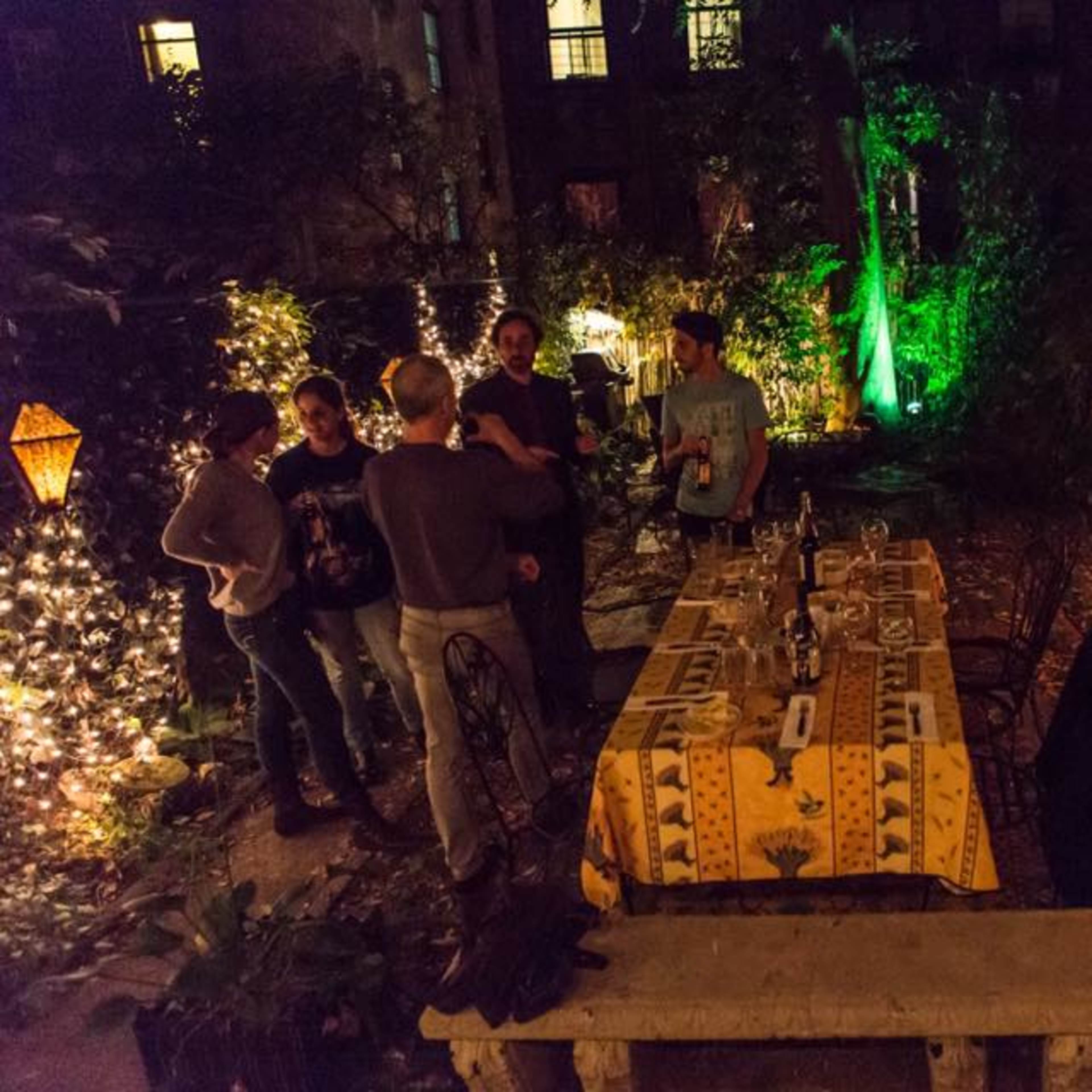 A group of people gathers around a table set in a garden adorned with string lights and lanterns.